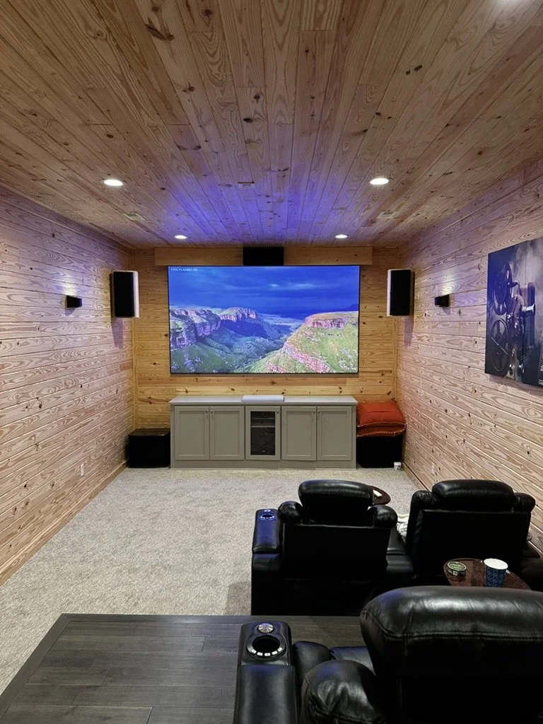 A cozy home theater room with wood-paneled walls and ceiling, a large flat-screen TV displaying a canyon landscape, black leather recliner chairs, and a side table with a cup and snack.