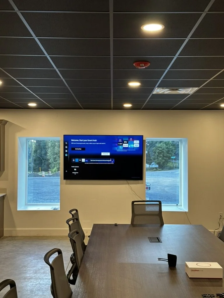A conference room with a long table, black office chairs, and a wall-mounted television screen displaying a setup menu. Two windows are on either side of the screen, showing an outdoor view of trees and a parking lot. The ceiling has recessed lights.