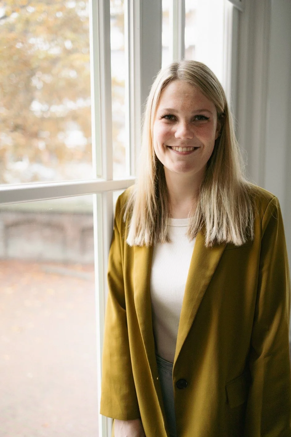 Vrouw met blond haar en gele blazer bij raam, glimlacht naar camera.
