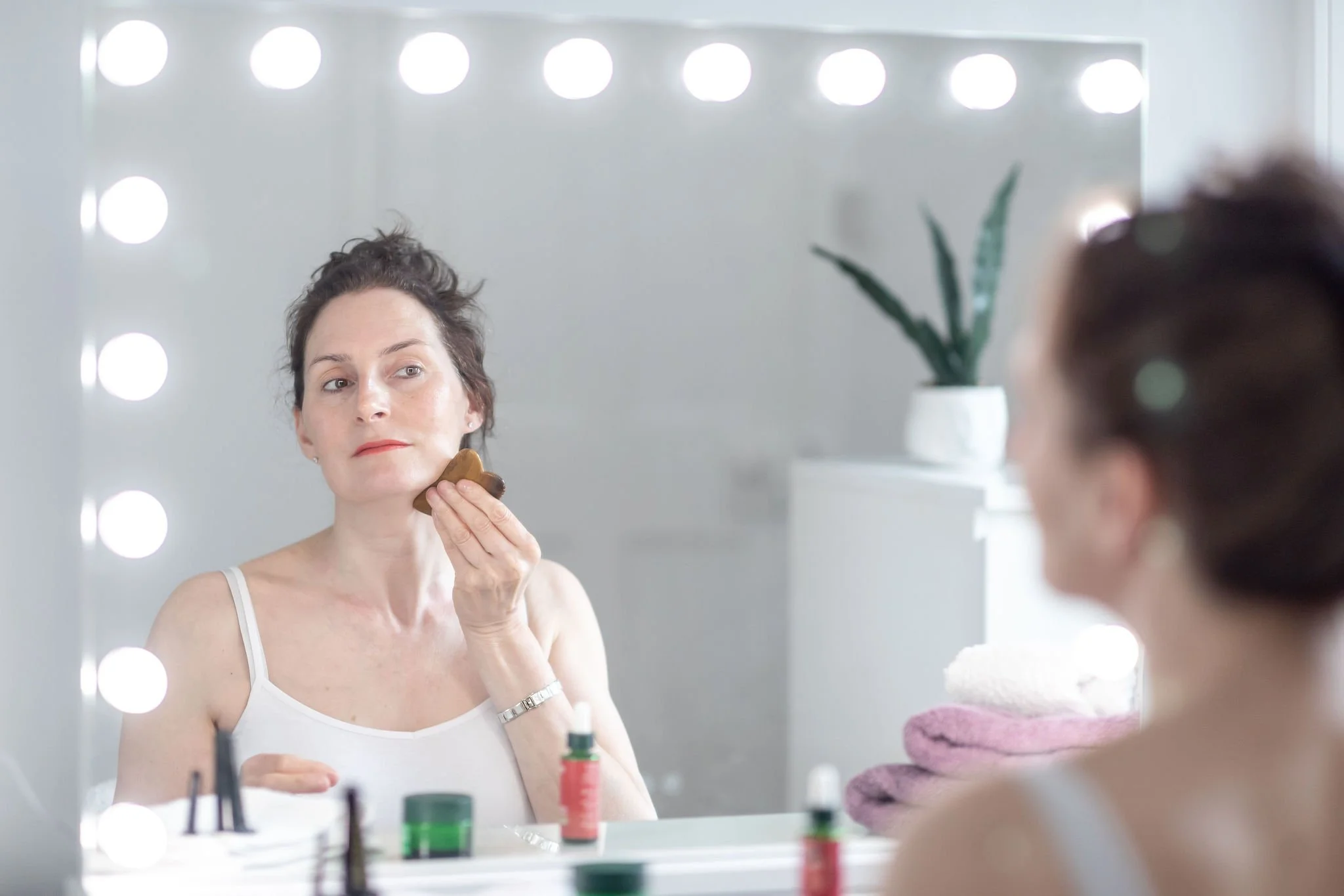 Woman applying makeup in front of a mirror with bright lights, holding a makeup sponge to her face, with various makeup products on the vanity and a potted plant in the background.