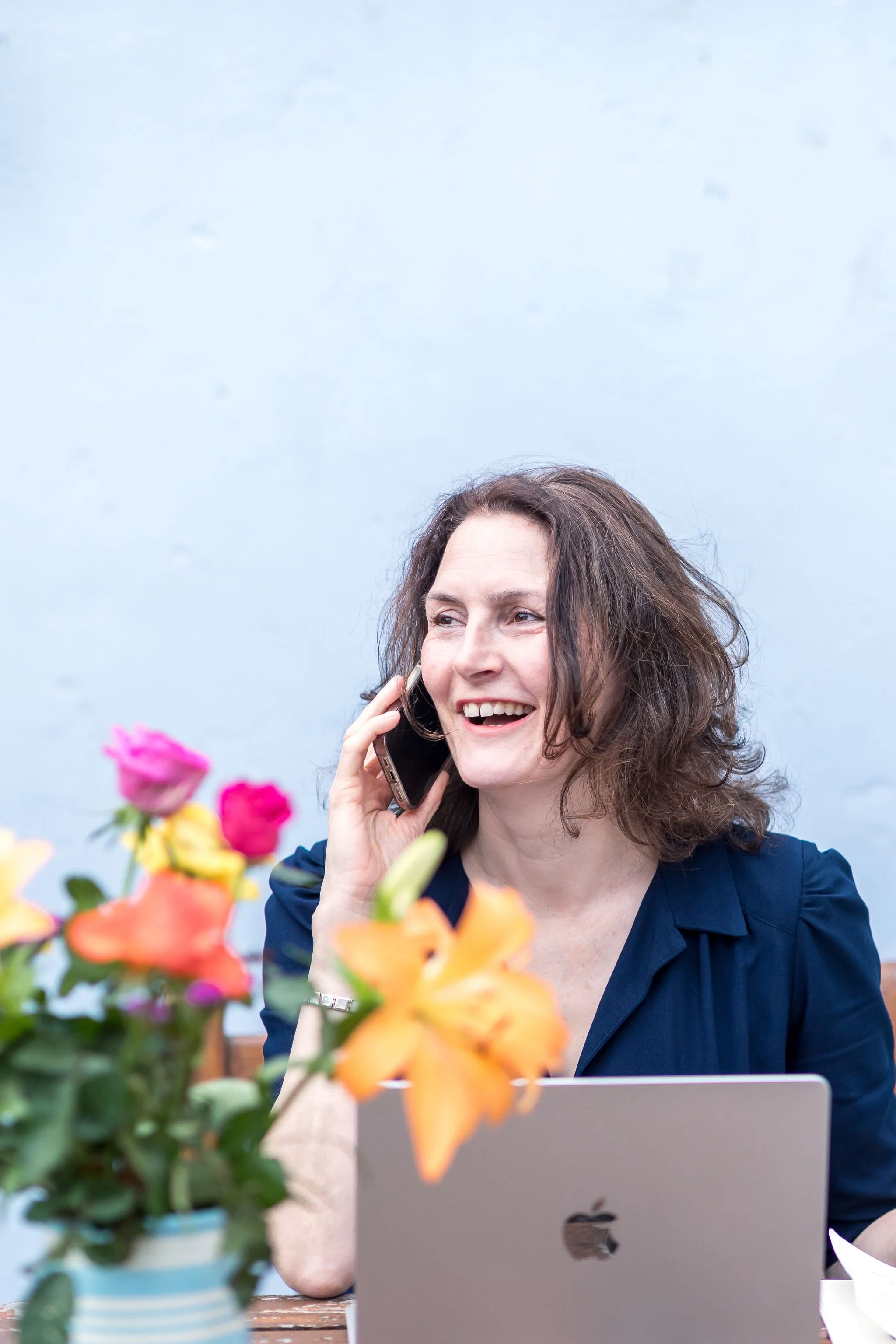 Five women sit around a dining table in a room decorated with framed paintings, opening presents and surrounded by gift items and beverages.