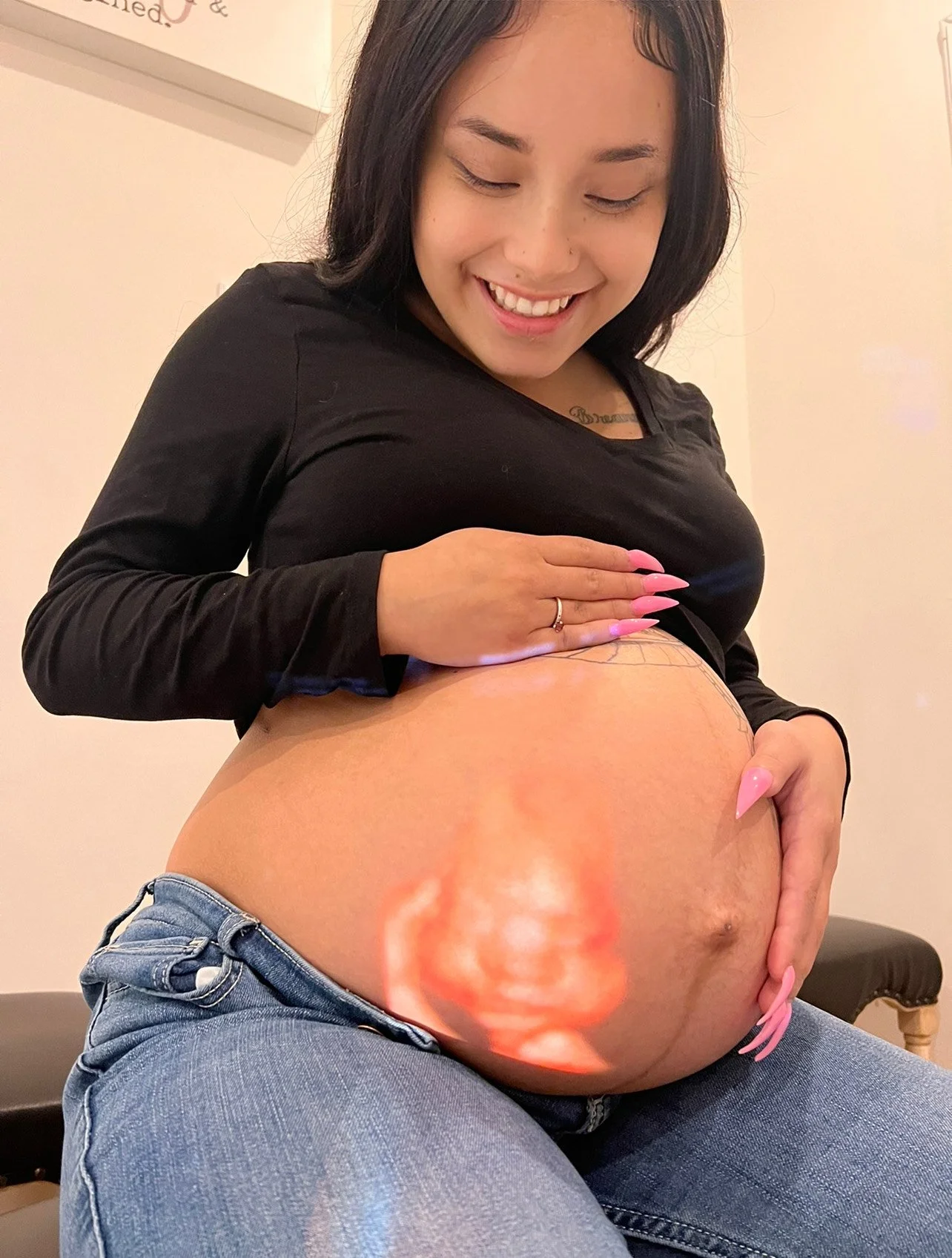 A woman with long dark hair, smiling and looking down, touches her large pregnant belly which has a digital ultrasound image of a baby's face on it. She is wearing a black top and blue jeans.