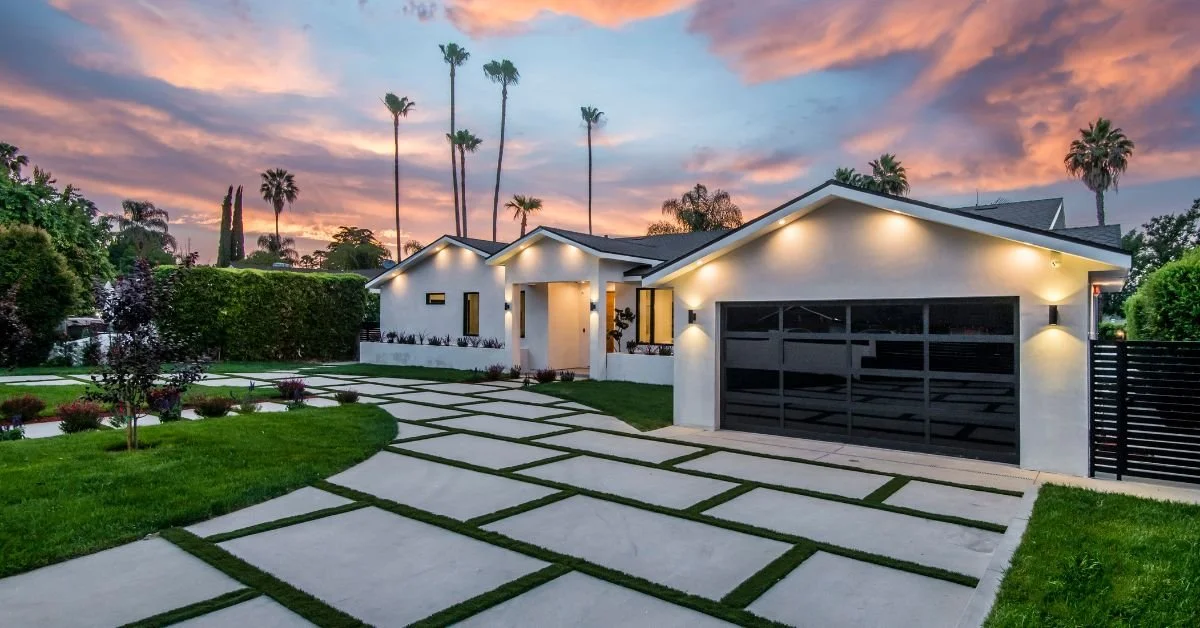 A white single-story modern house with a black garage door and a concrete driveway divided by grass under a pink sunset sky.