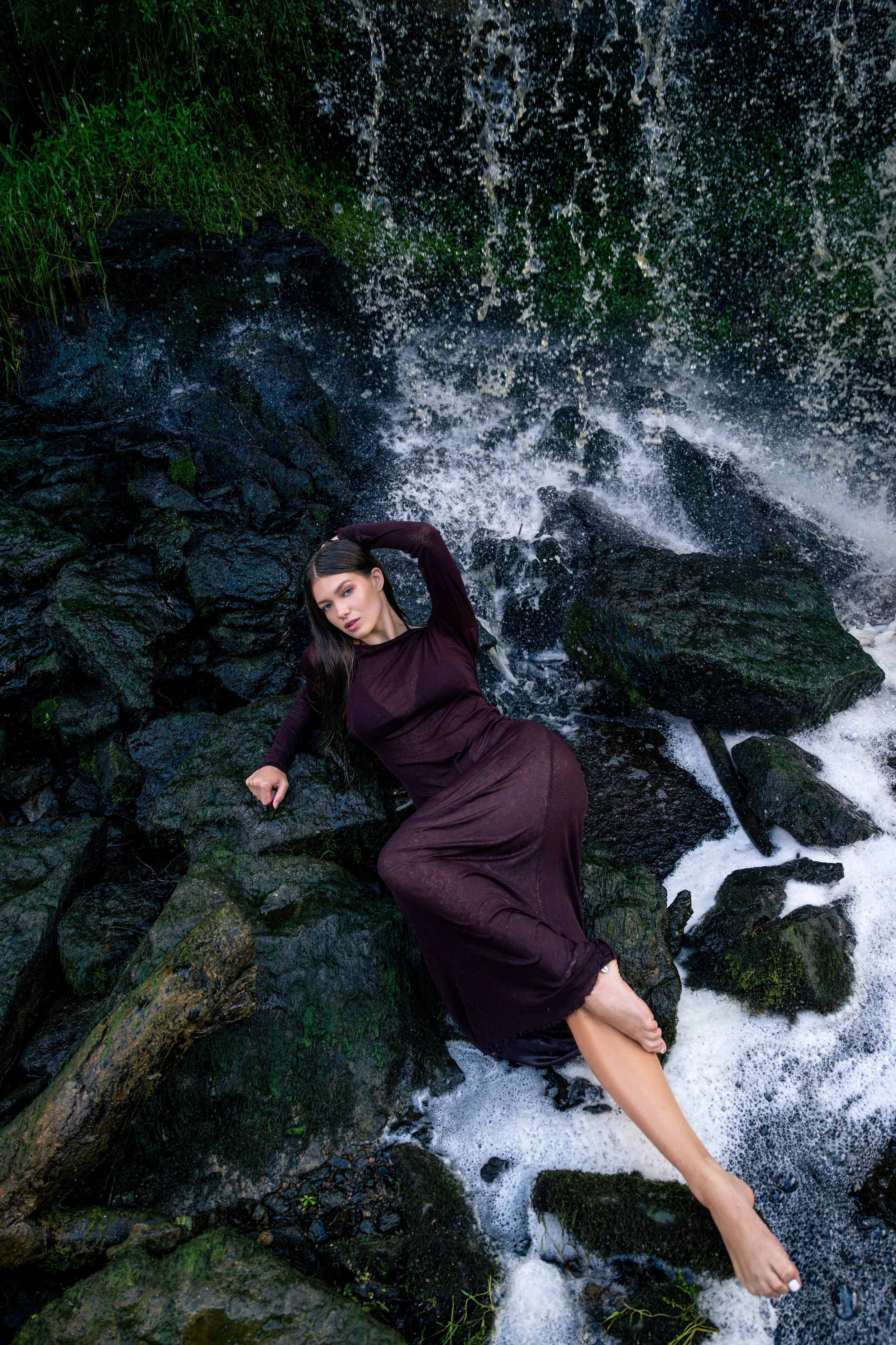 A woman with long dark hair is lying on wet rocks in a stream with water splashing behind her. She is wearing a long, dark dress and has one hand behind her head while the other rests on the rocks. Her eyes are partially closed, and she looks directl