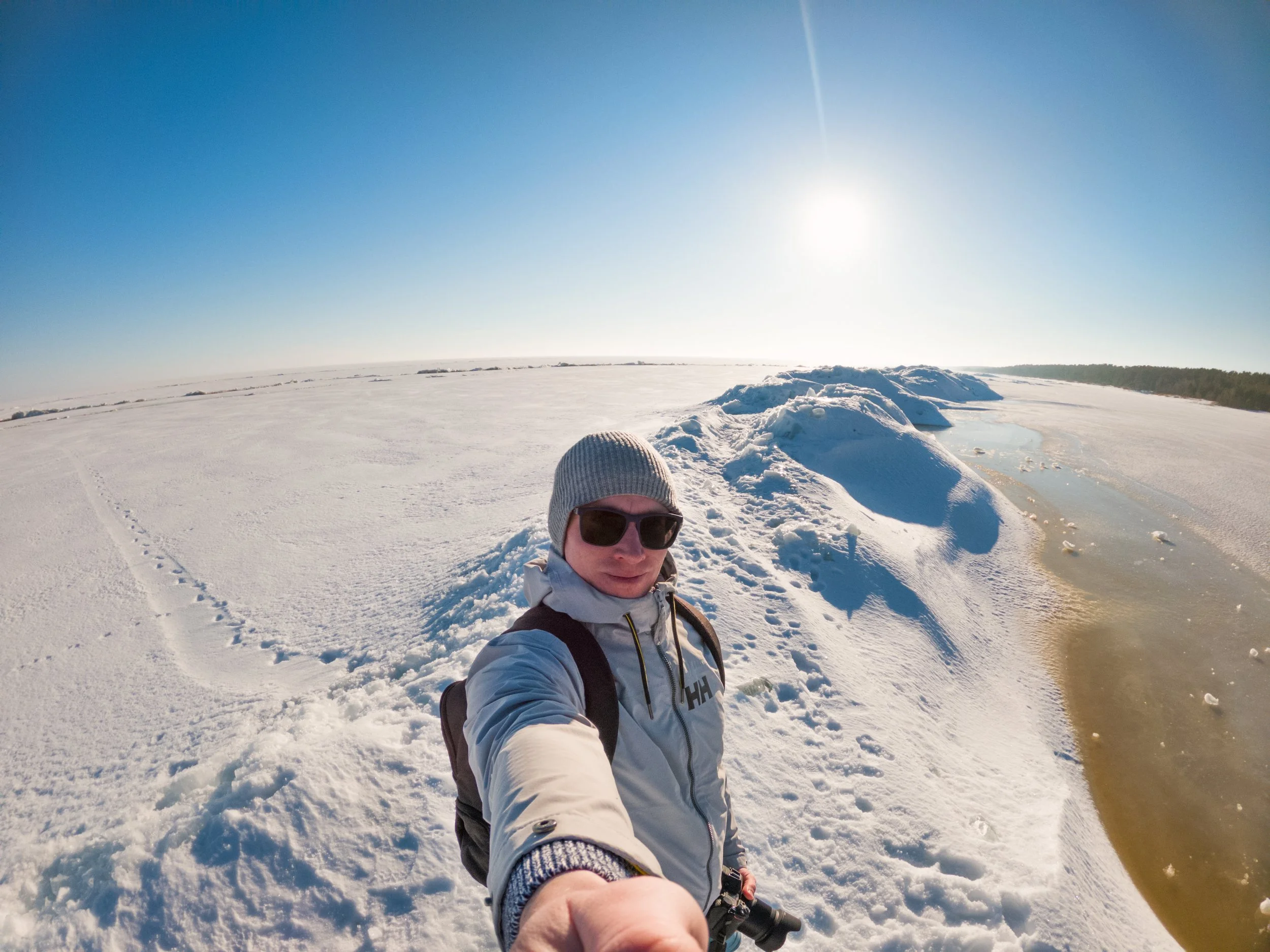 A person in winter gear, wearing sunglasses, a beanie, and a backpack, takes a selfie on a snow-covered landscape near a frozen body of water with ice mounds and a bright sun overhead.