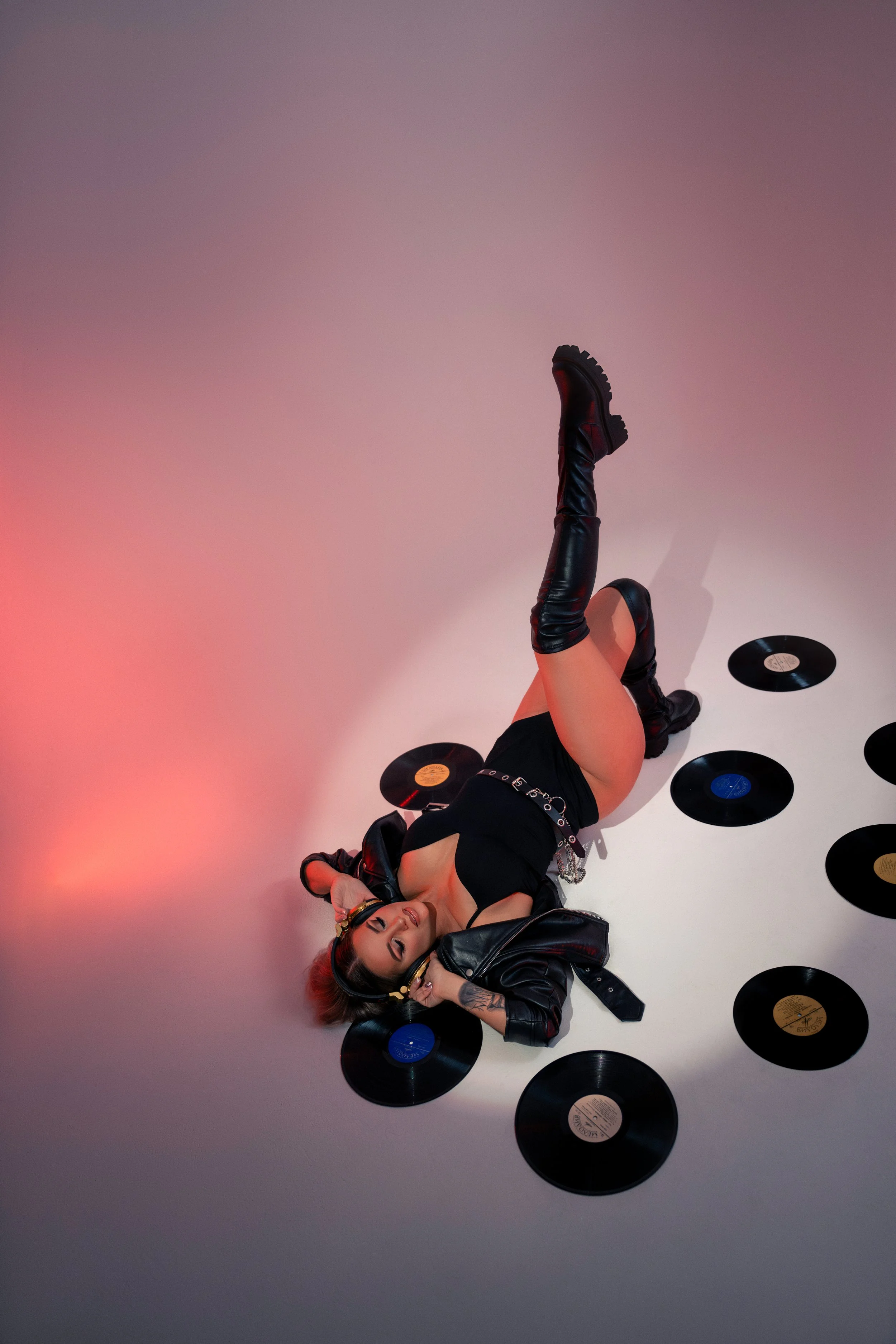 A woman with red hair and tattoos lying on the floor surrounded by vinyl records, wearing black boots, a black dress, and a leather jacket, with her eyes closed and hands near her face, in a room with colorful lighting.