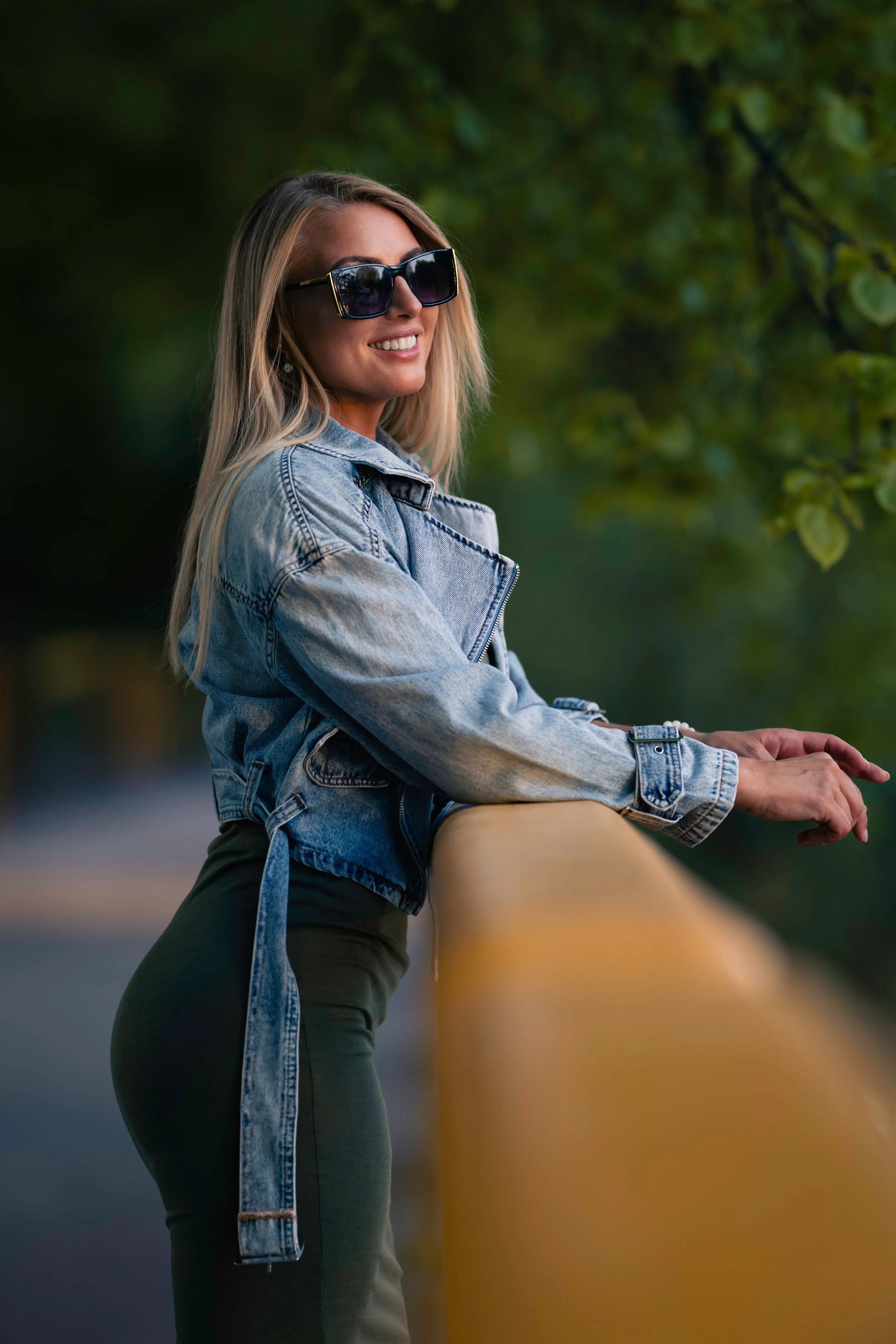 A smiling woman with blonde hair, wearing large dark sunglasses, a denim jacket, and black pants, leaning on a yellow railing outdoors, with blurred green foliage in the background.