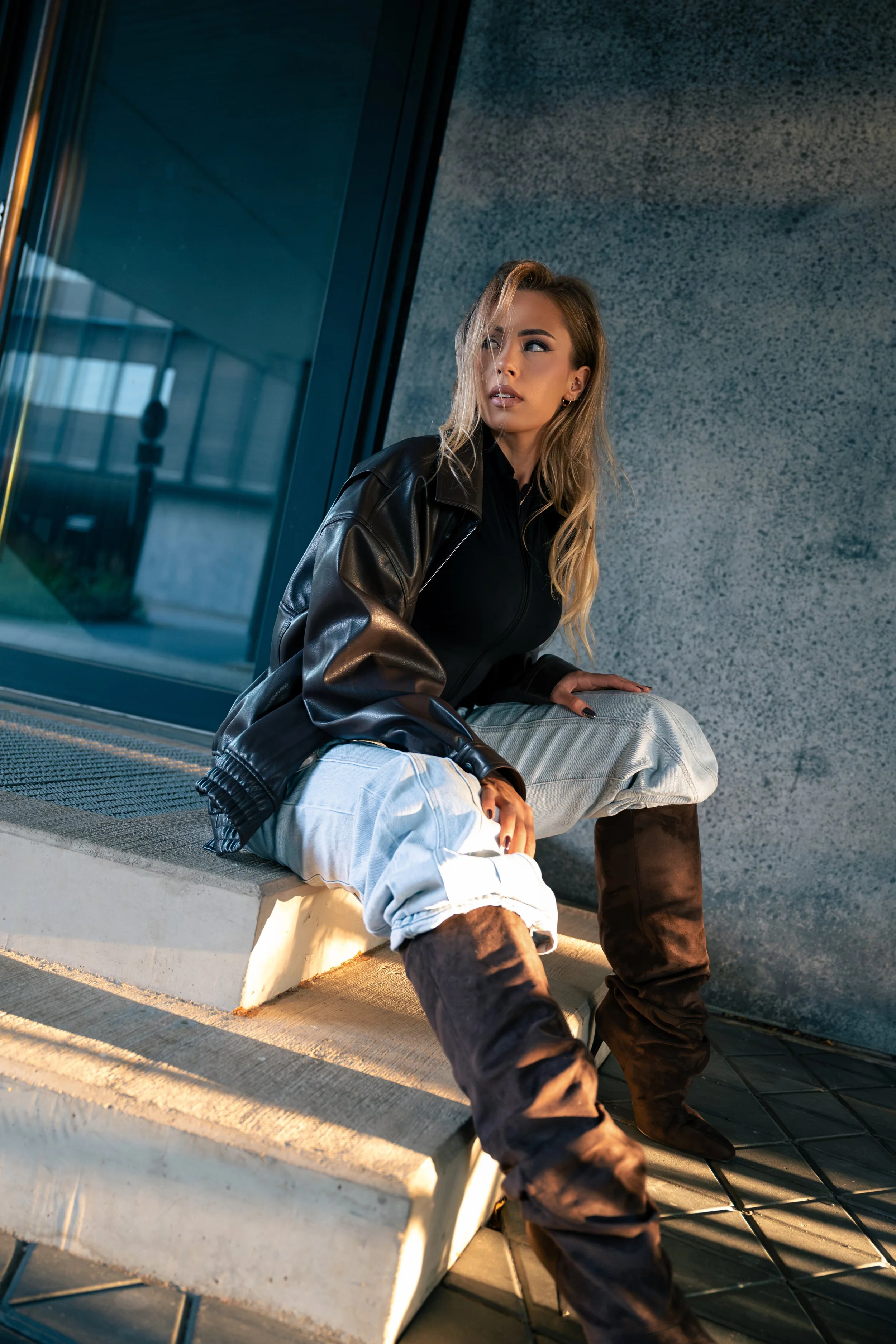 A young woman with blonde hair, wearing a black leather jacket and white pants, sitting on a bench outside near a modern building with large glass windows and blue-tinted panels, lit by warm sunlight.