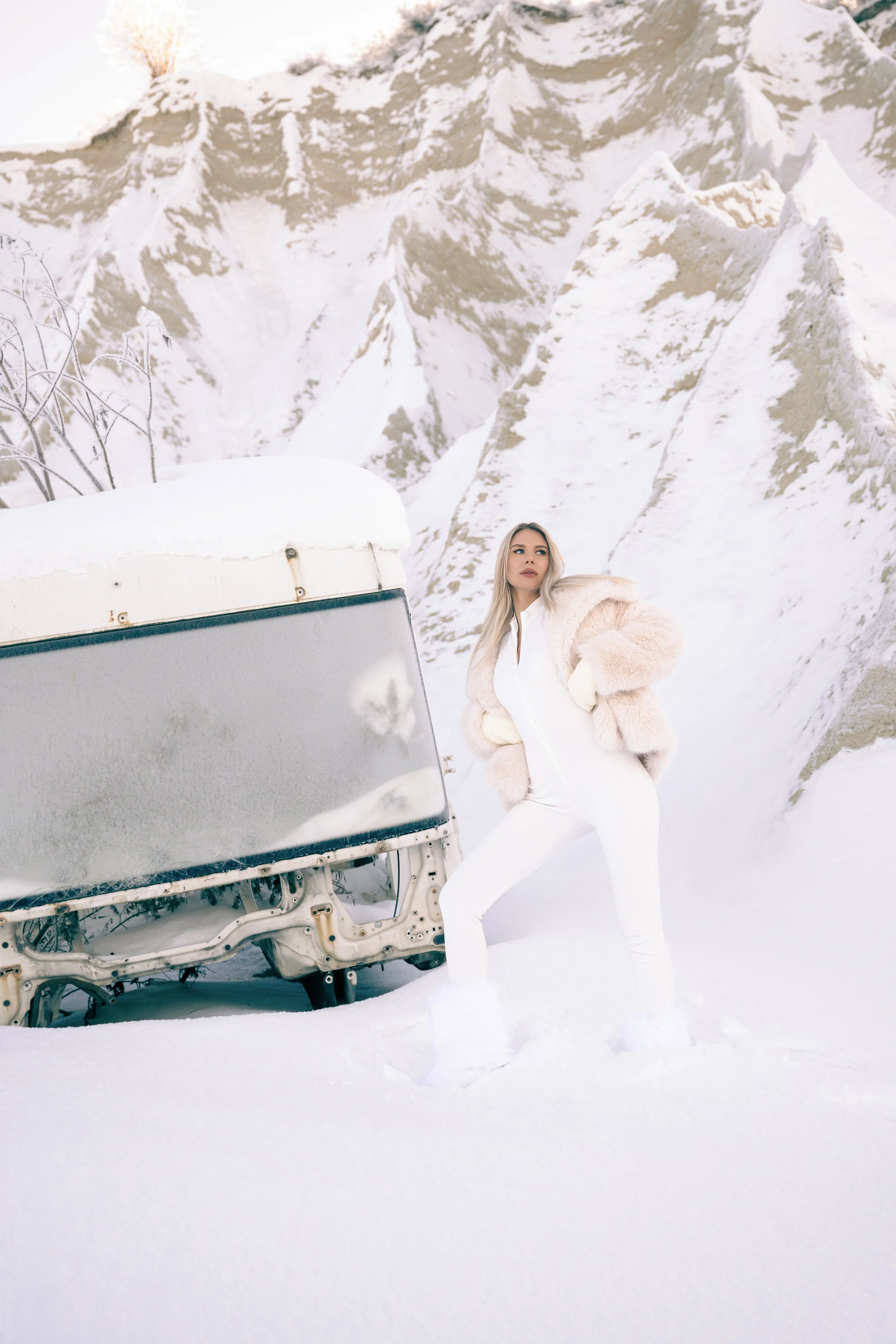 A woman in a white outfit and beige fur coat stands in snow next to an abandoned, snow-covered vehicle with mountains in the background.