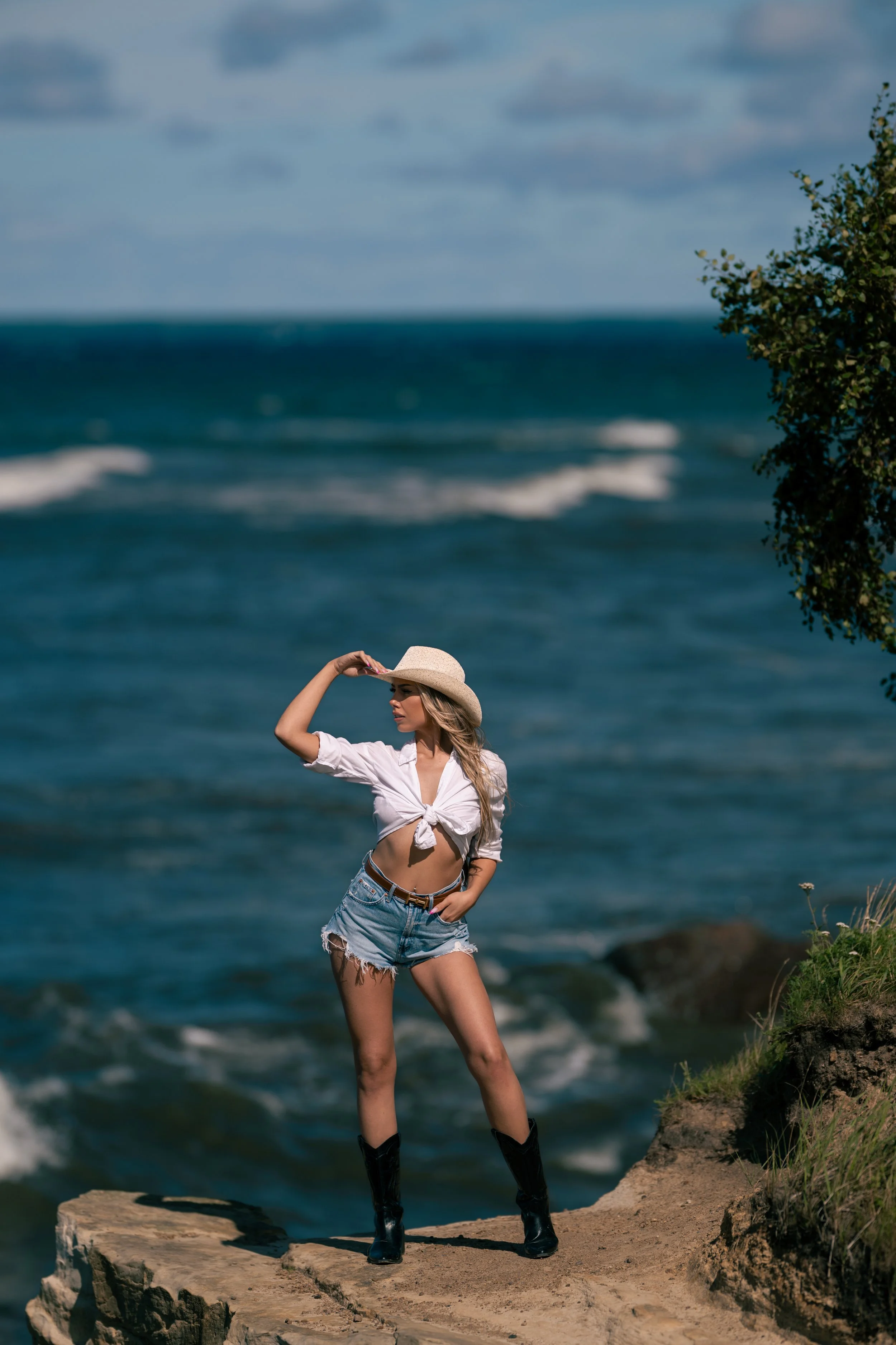 A young woman in a white knotted shirt, denim shorts, and black boots standing on a rocky ledge near the ocean, wearing a wide-brimmed hat, holding it with one hand, with the sea and cloudy sky in the background.