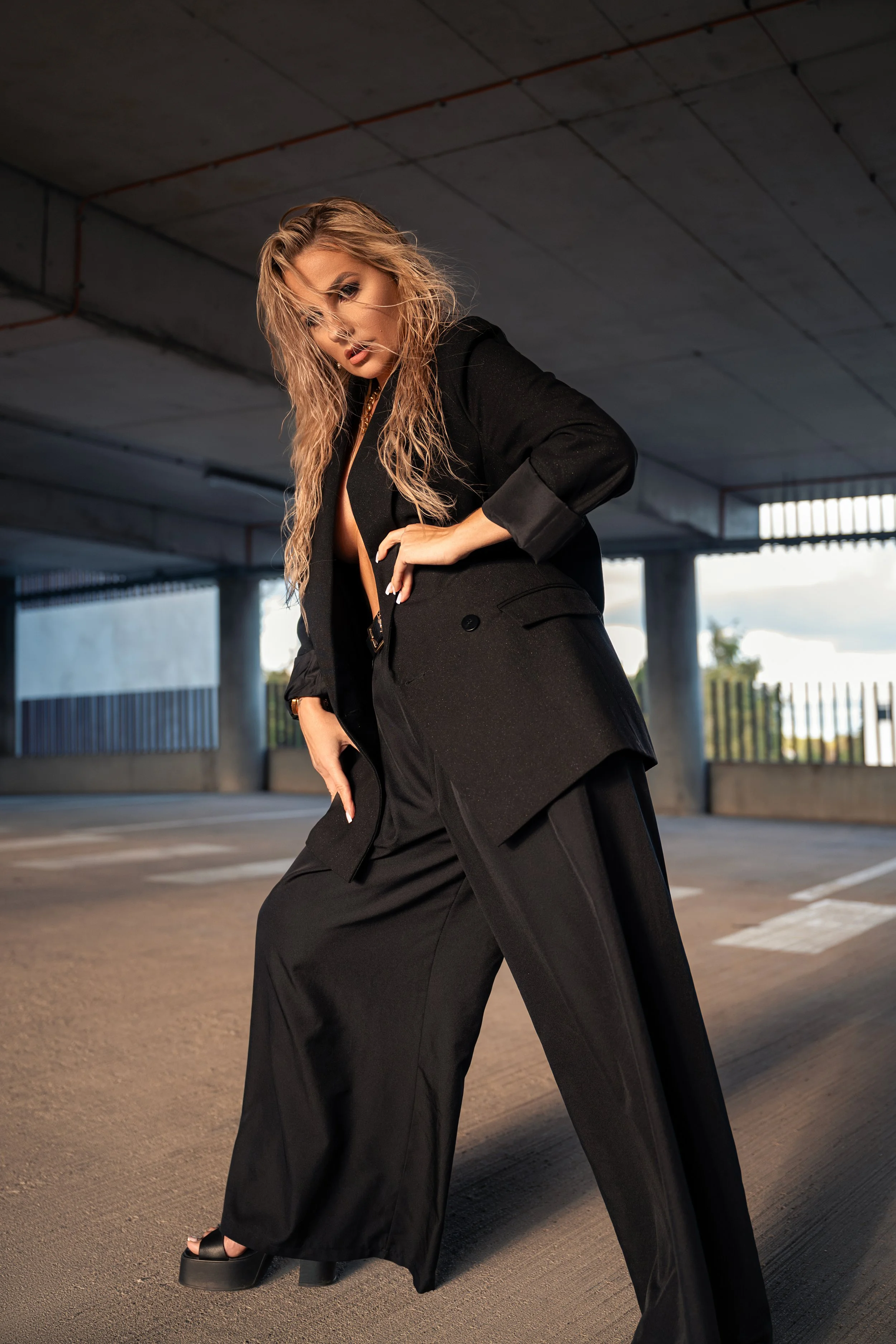 A woman with wet, wavy blonde hair, wearing a black blazer and black wide-leg pants, standing in an empty parking garage during daylight, looking at the camera with a serious expression.