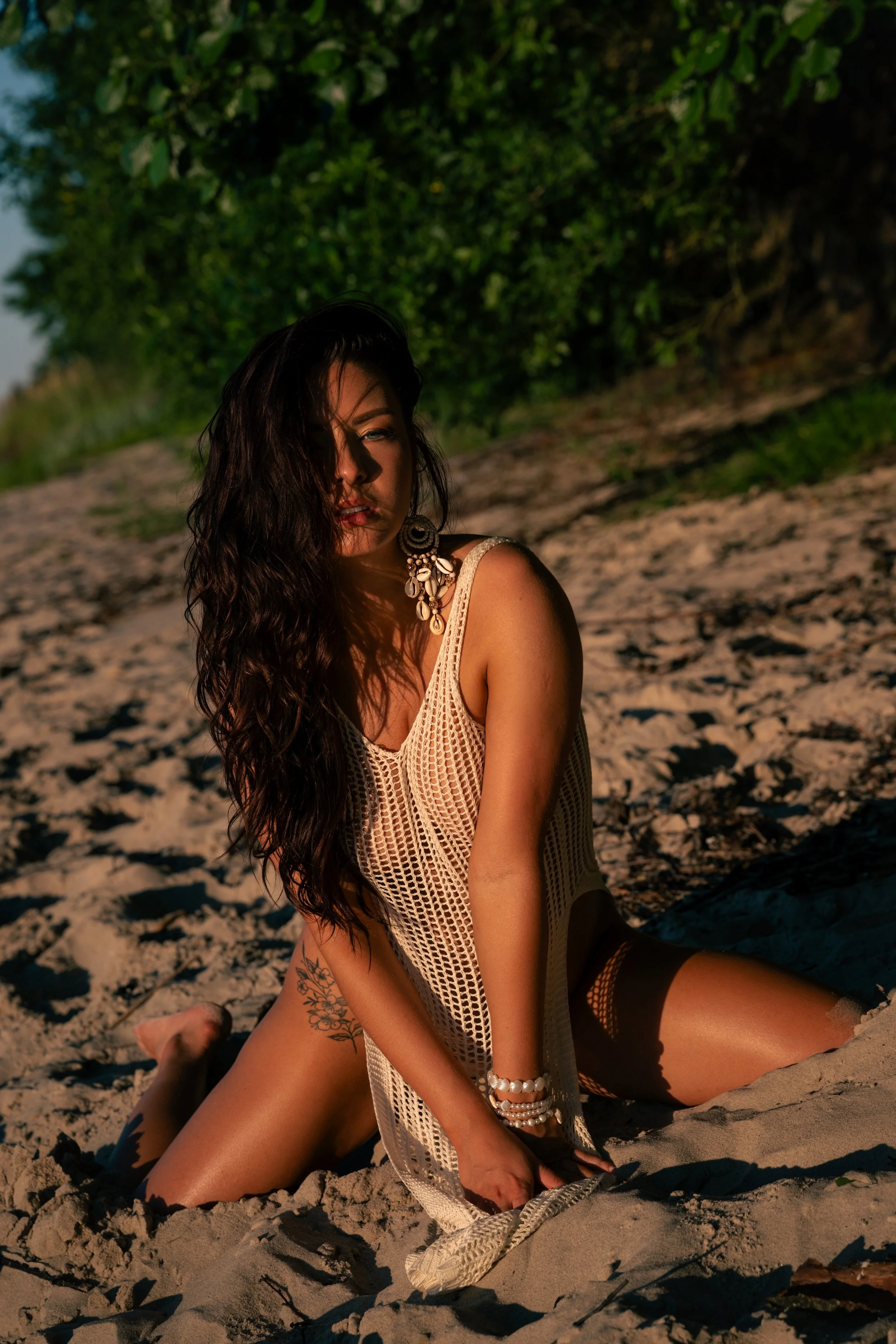 A woman with long dark hair and tattoos is sitting on a sandy beach during sunset, wearing a beige crochet top and jewelry, with a lush green background.