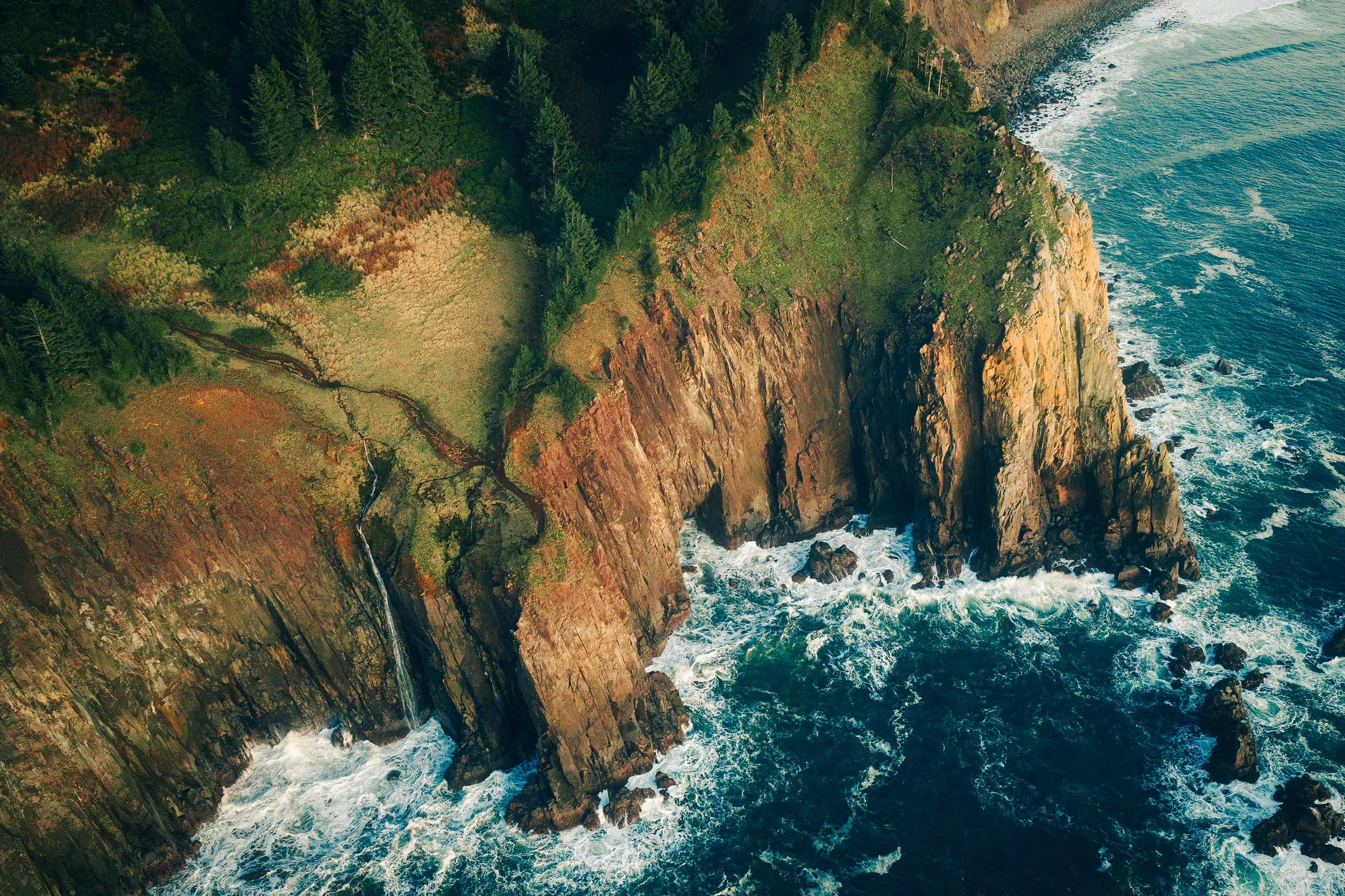 Aerial view of rugged coastline with green forest, rocky cliffs, and ocean waves crashing at the shore.