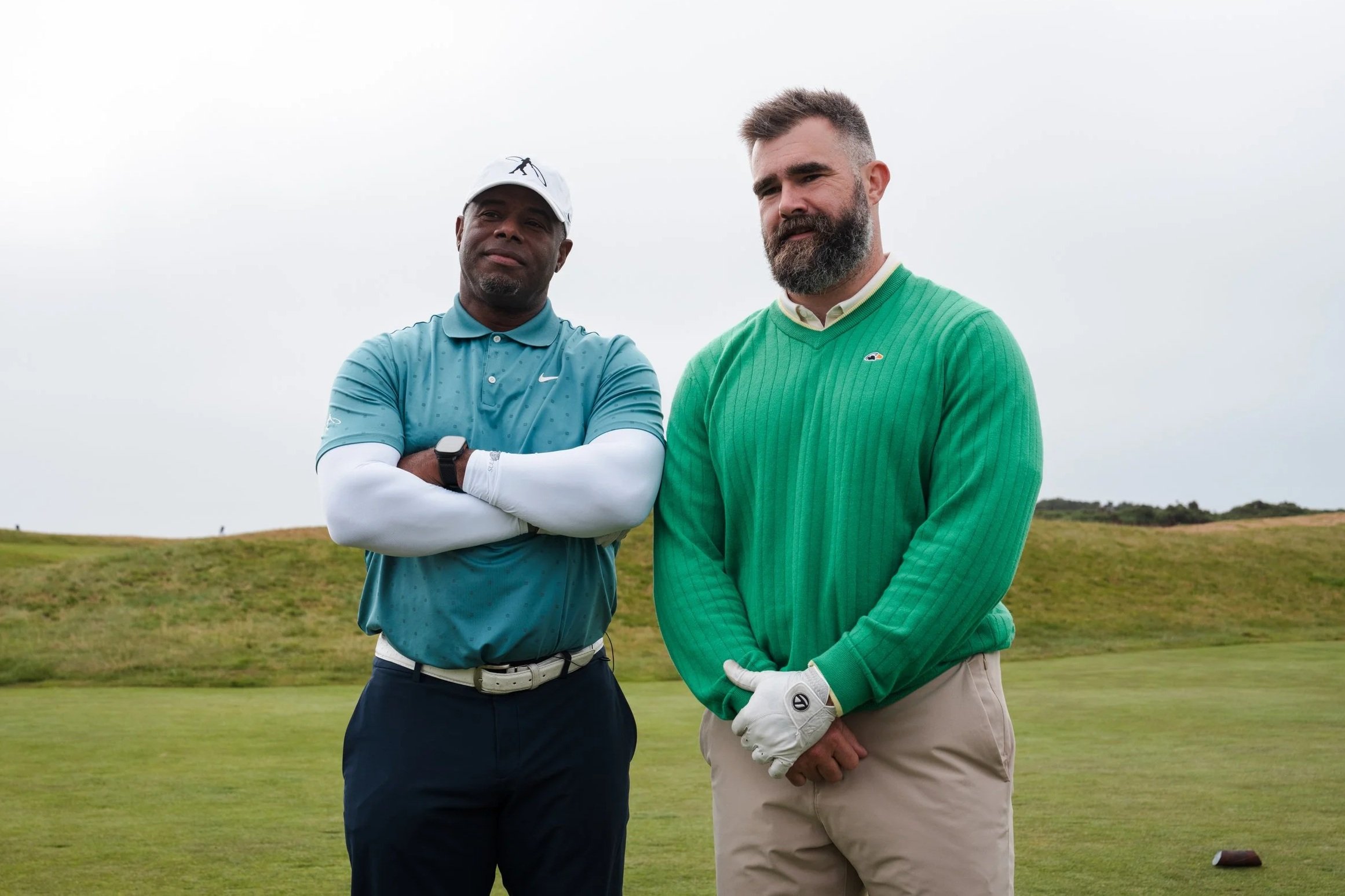 Two men standing outdoors on a golf course, one wearing a light blue polo shirt and a white cap, the other wearing a green sweater and beige pants, both posing with arms crossed in a cloudy sky setting.