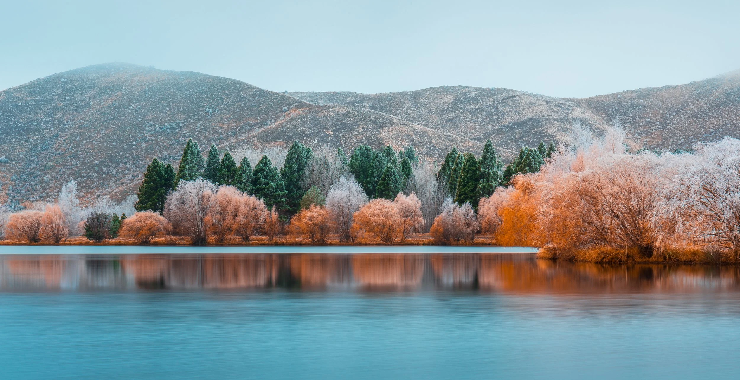 Landscape with a calm lake in the foreground, surrounded by trees with autumn-colored foliage, and mountains in the background under a clear sky.