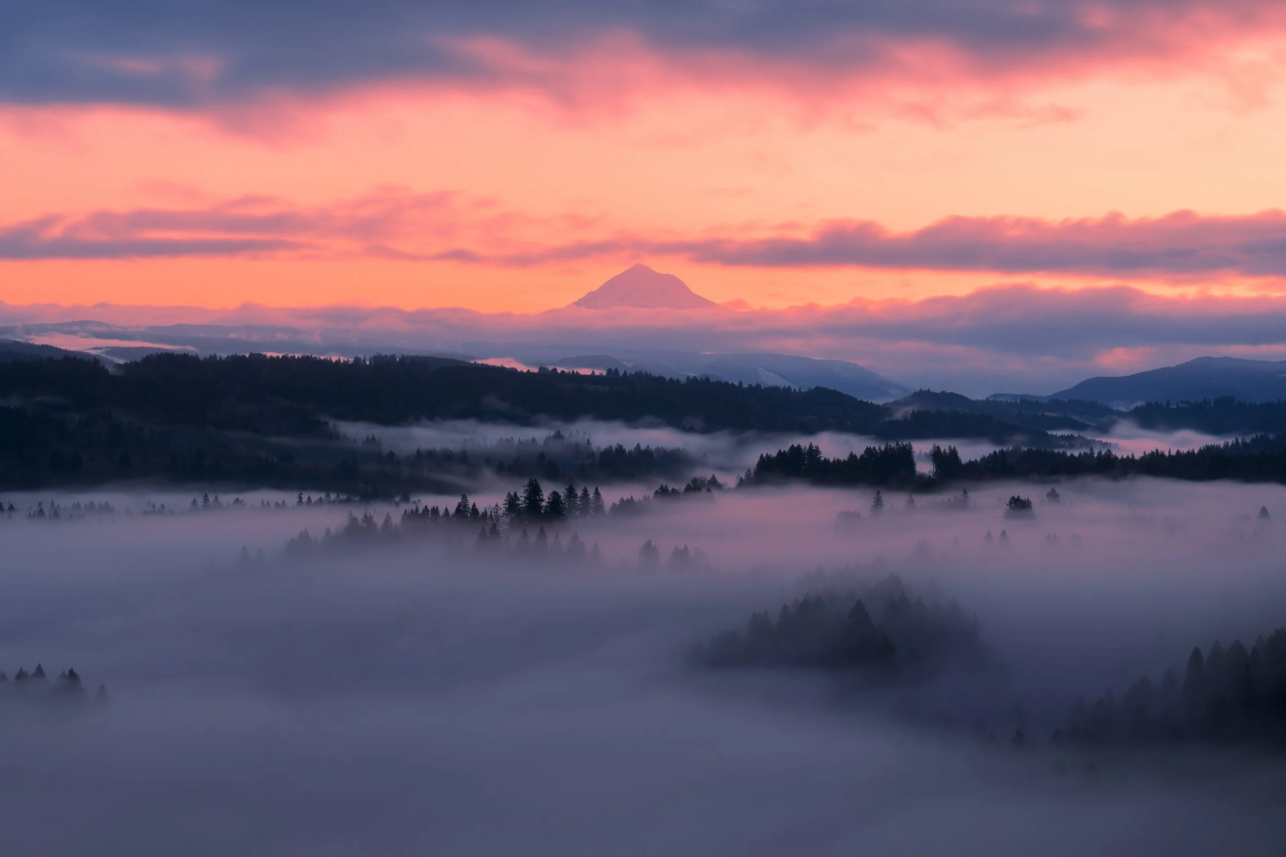 Sunset over a misty landscape with layers of trees and hills, with a mountain in the distance, and colorful clouds in the sky.