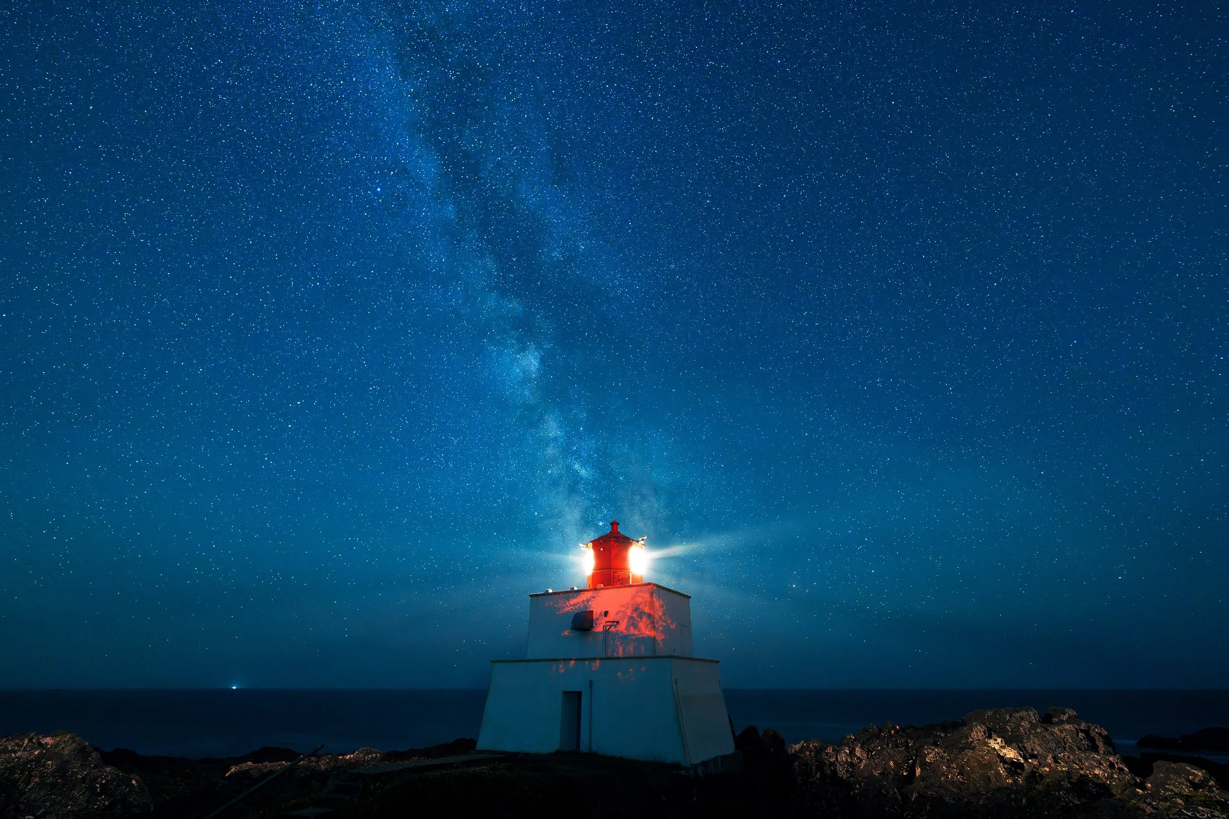 Night sky filled with stars and the Milky Way galaxy shining above a lighthouse on rocky coastline