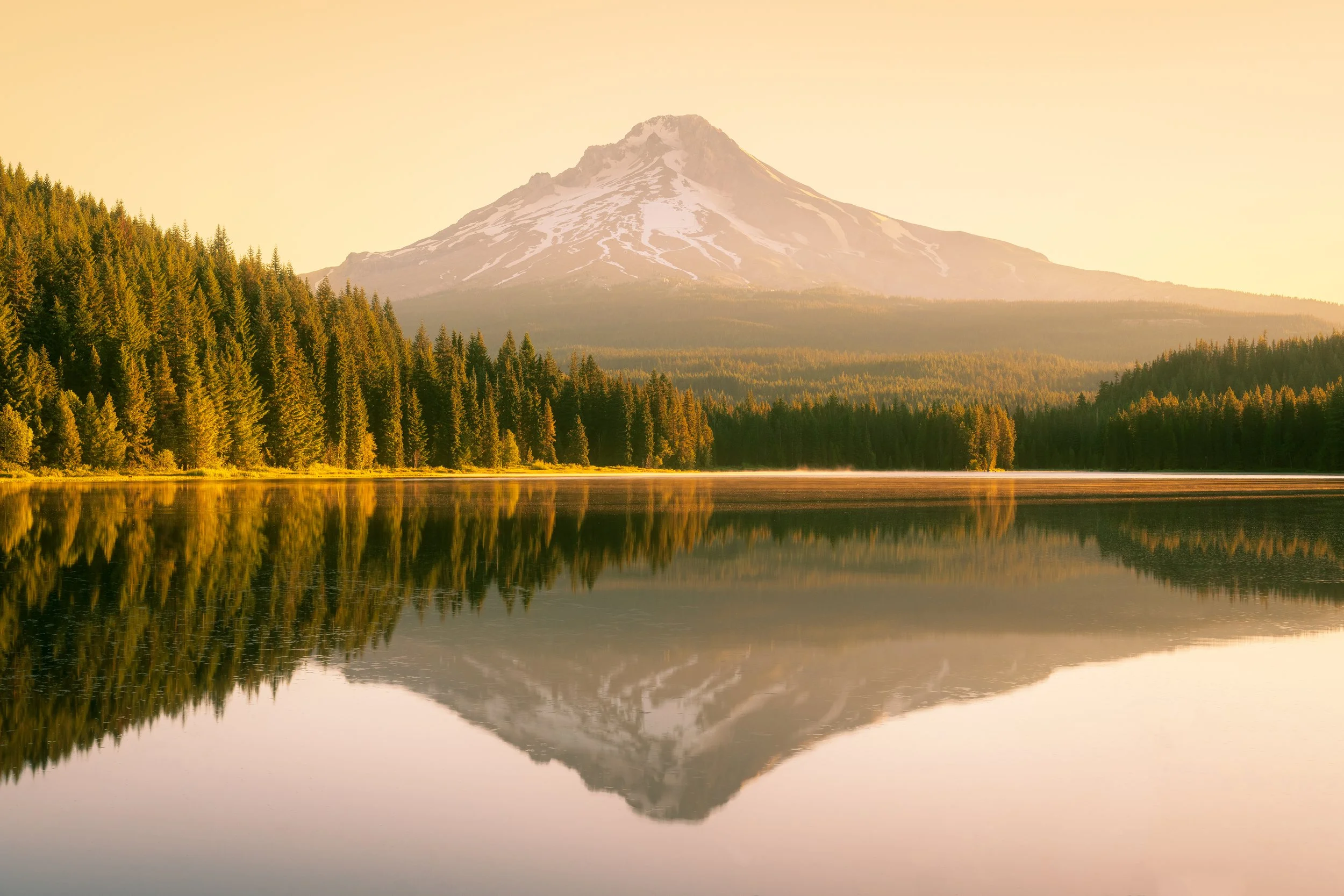 A mountain with snow on its peak reflected in a calm lake surrounded by a dense forest at sunset.