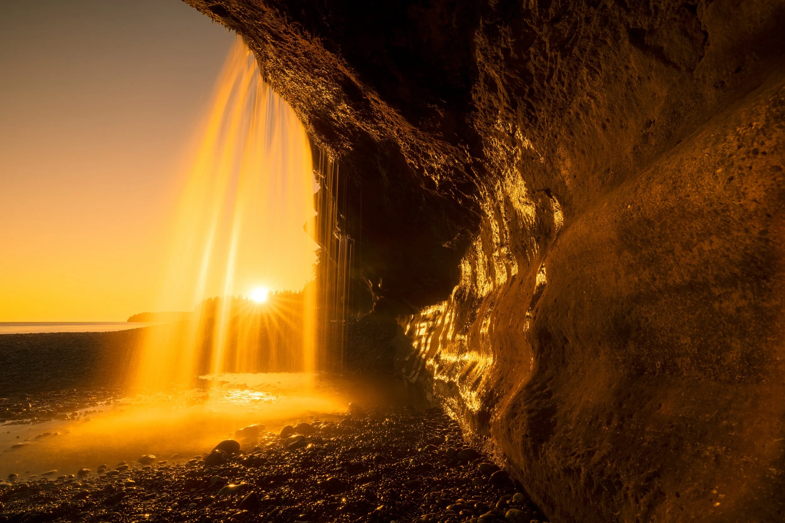 A golden sunset viewed from underneath a waterfall on a rocky beach.