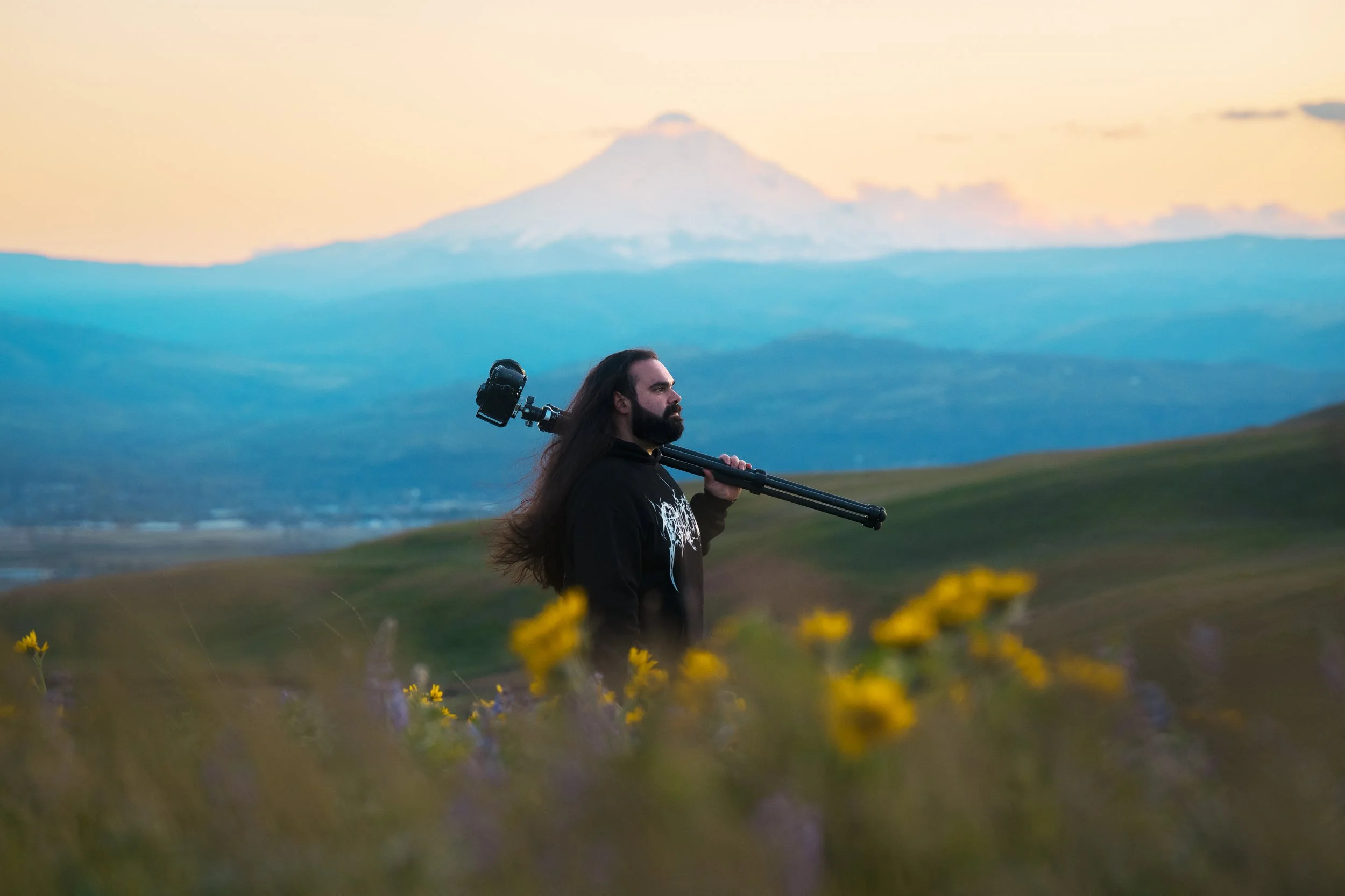 A man with long dark hair and a beard carrying a camera tripod on his shoulder, standing in a field of yellow and purple flowers with a mountain in the background during sunrise or sunset.