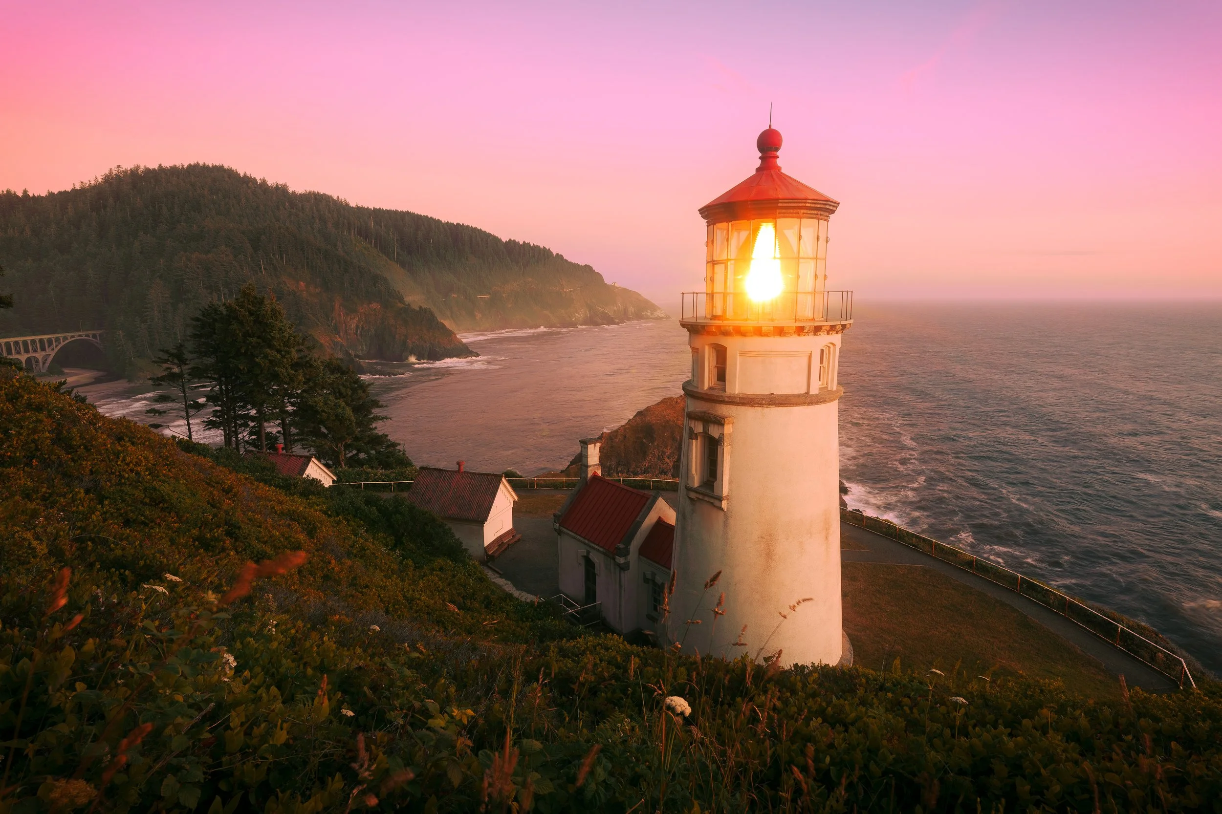 Lighthouse at sunset on a coastal cliff with a pink and purple sky, overlooking the ocean with waves and a forested hillside in the background.