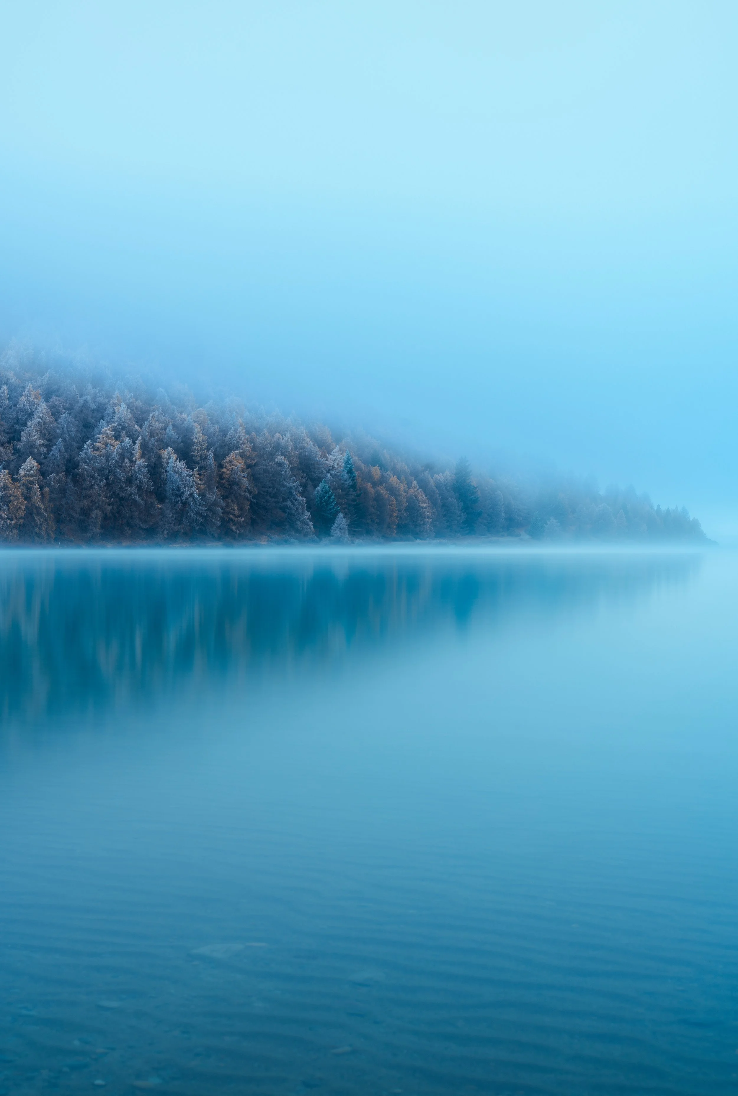 A calm, blue lake surrounded by snowy trees and misty mountains in the background.