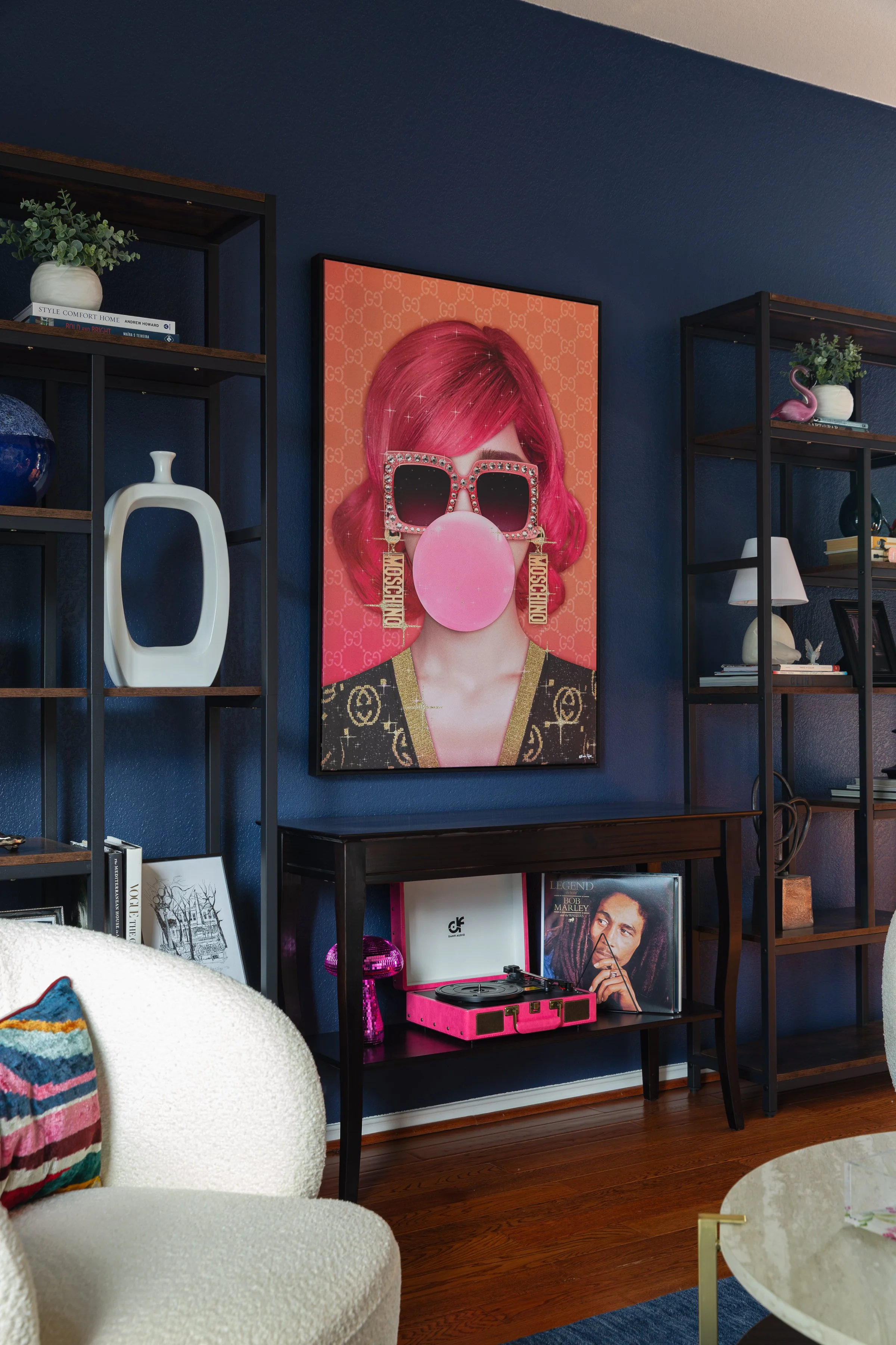 Living room with dark blue walls, featuring a large colorful artwork of a person with pink hair and bubblegum, surrounded by decorative shelves. A record player and vinyl records sit beneath the artwork on a dark console. A white plush chair and roun