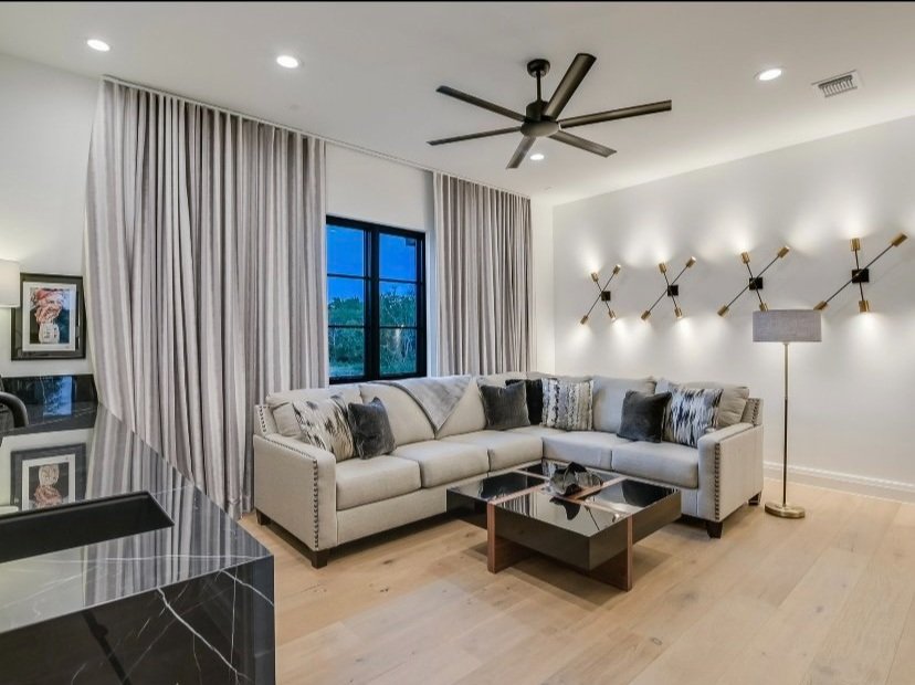 Modern living room with gray sectional sofa, decorative pillows, geometric wall lights, ceiling fan, wooden floor, black window, beige curtains, and a reflective coffee table.