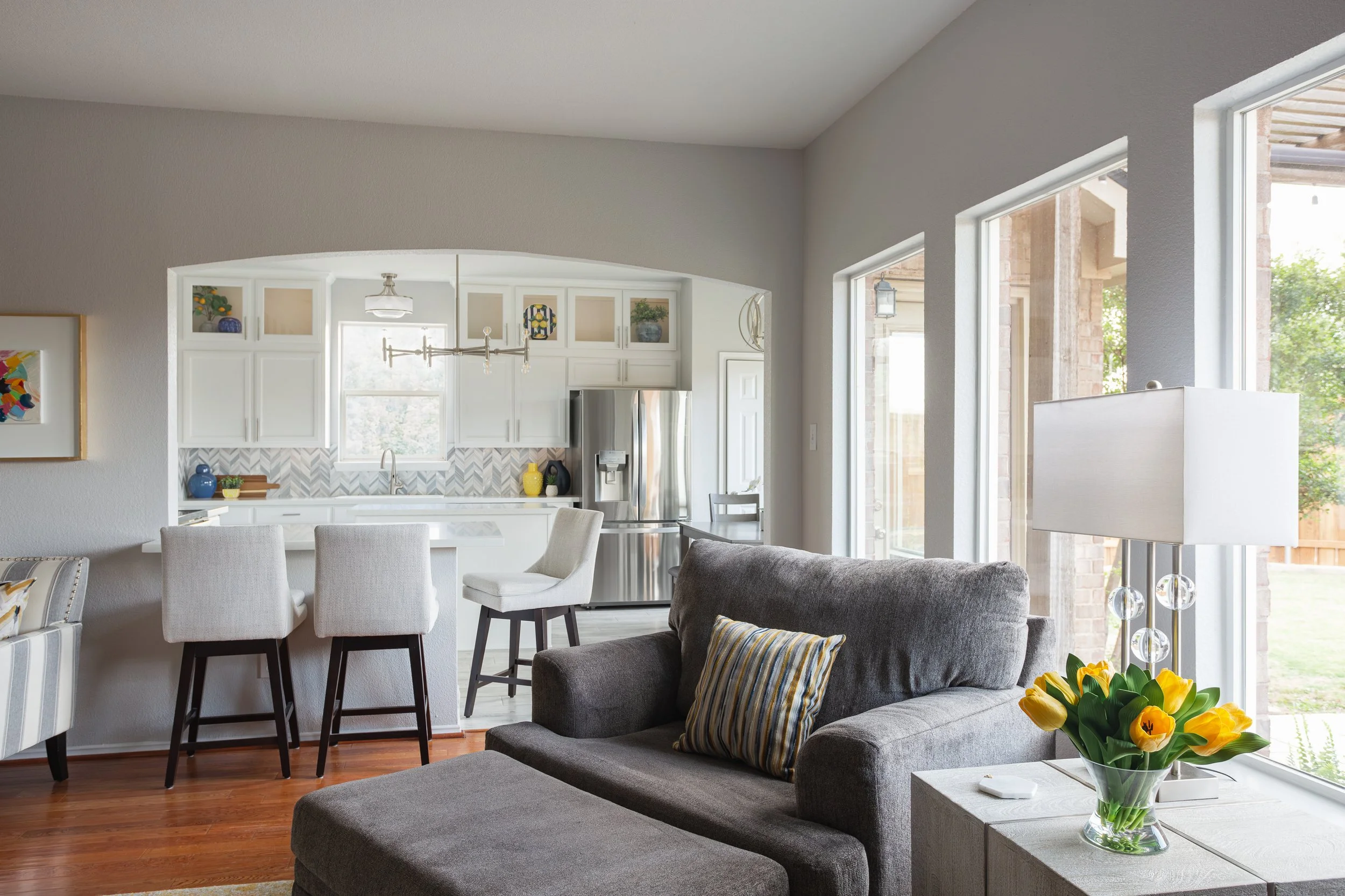Modern living room and kitchen with gray sofa, yellow tulips in a vase, bar stools, white kitchen cabinets, stainless steel appliances, and large windows.