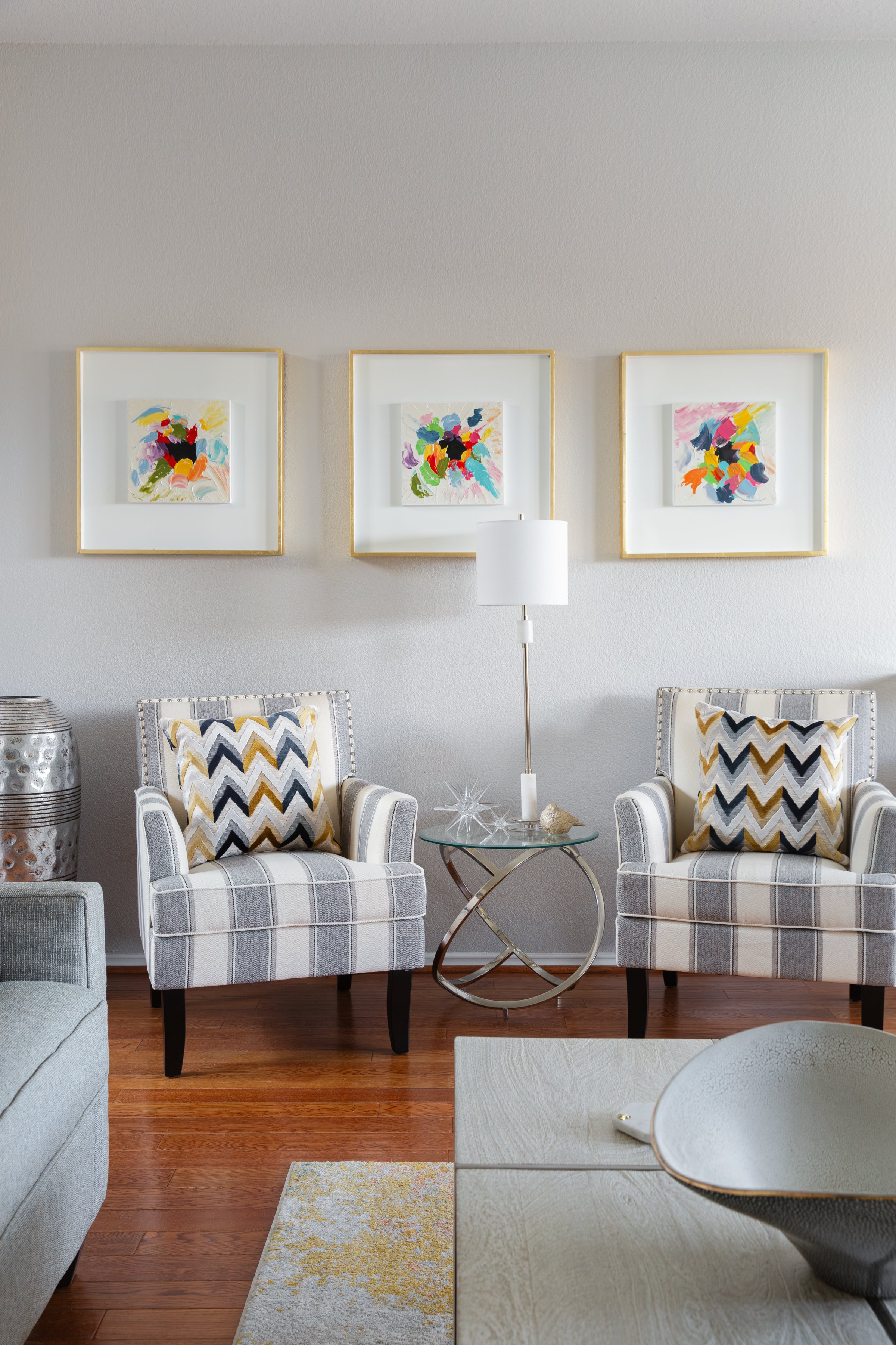 Living room with two striped armchairs, decorative pillows, round glass side table, and abstract wall art.