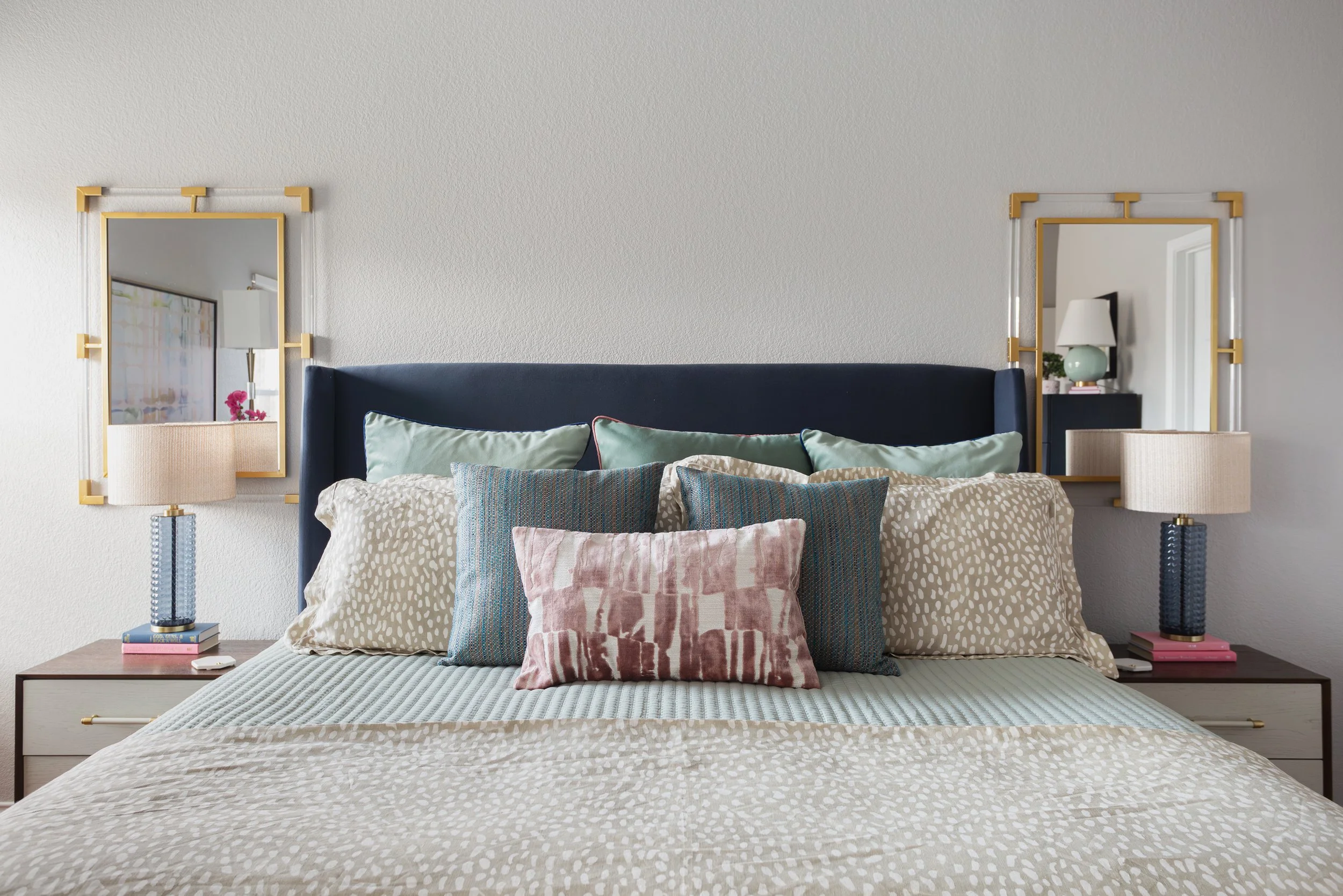 A neatly arranged bedroom with a blue upholstered headboard, patterned pillows, and a neatly made bed. Two side tables hold lamps and books, and two rectangular mirrors with gold accents are hung above the tables.