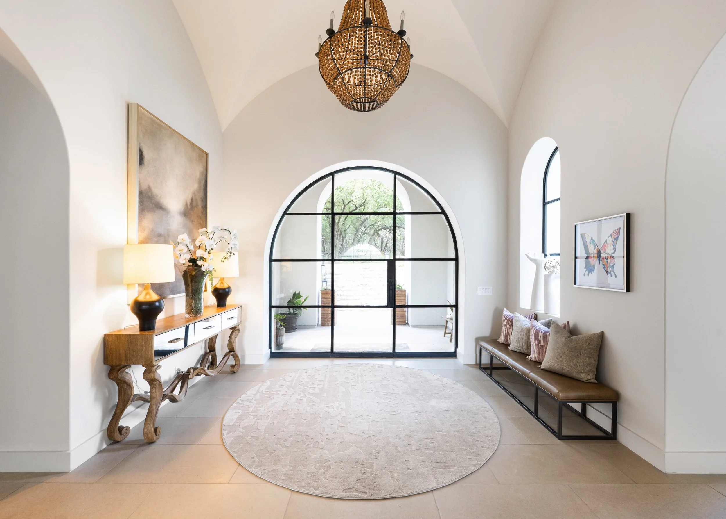 Elegant entryway with high arched ceiling, large window, round beige rug, wooden console table with lamps and flowers, bench with decorative pillows, and artwork on walls.