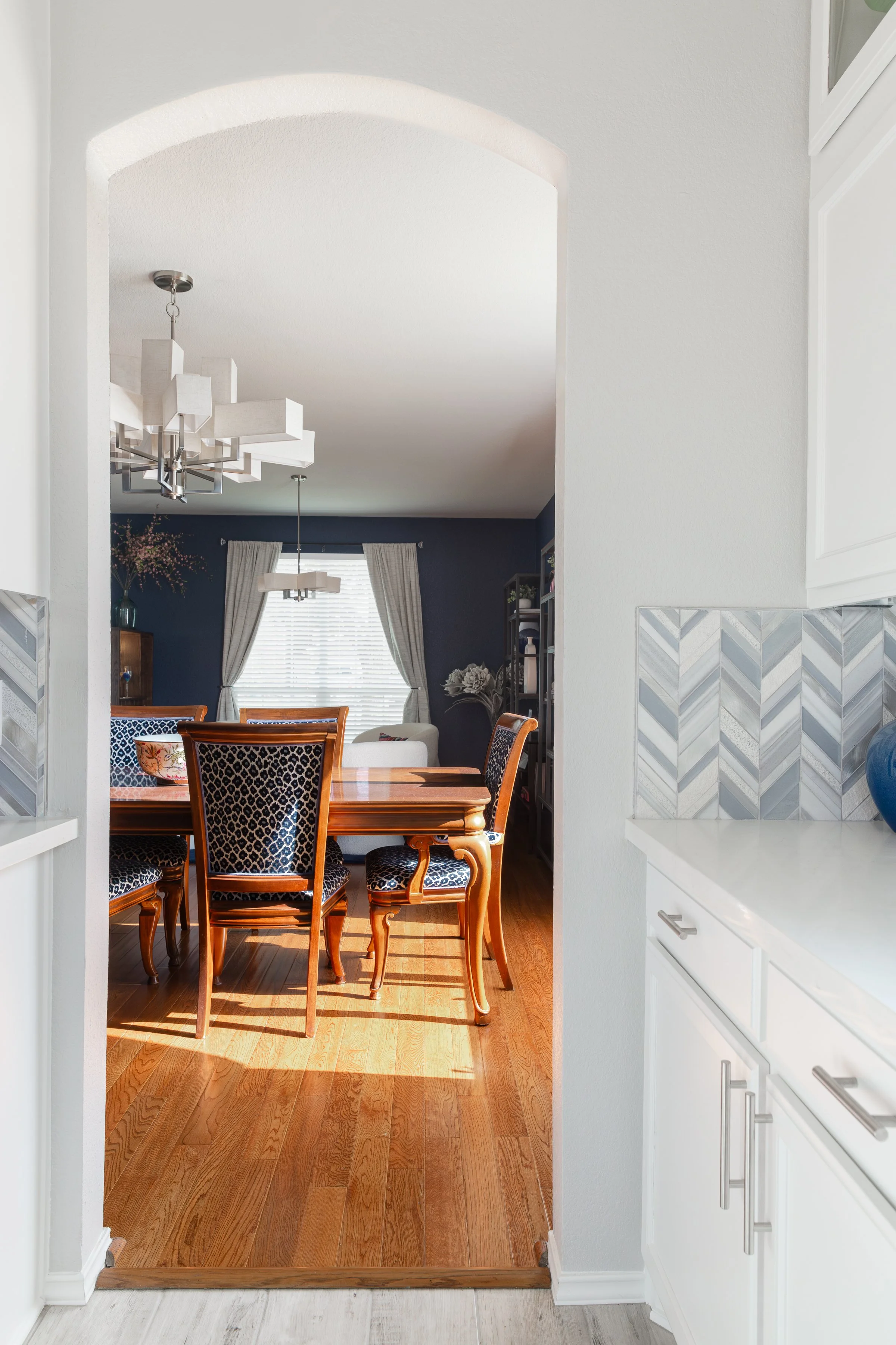 Elegantly decorated dining room with arched entryway, wooden table and chairs, blue upholstered seats, geometric tile backsplash, and modern chandelier.