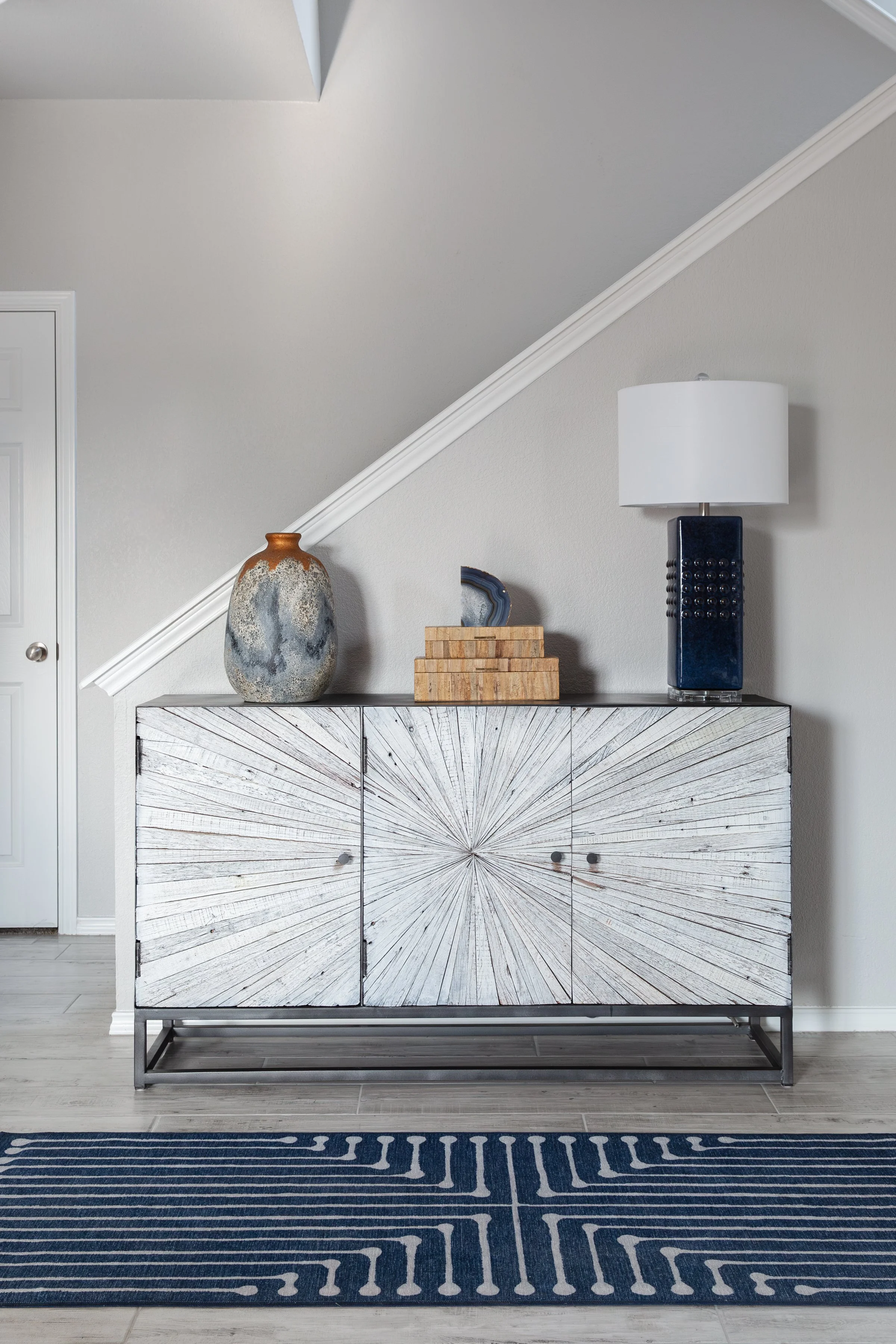 Interior design with a white cabinet featuring starburst pattern, a decorative blue and orange vase, a stack of wooden blocks, a tall lamp on top of the cabinet, a blue and white patterned rug, and a gray wall with a staircase.
