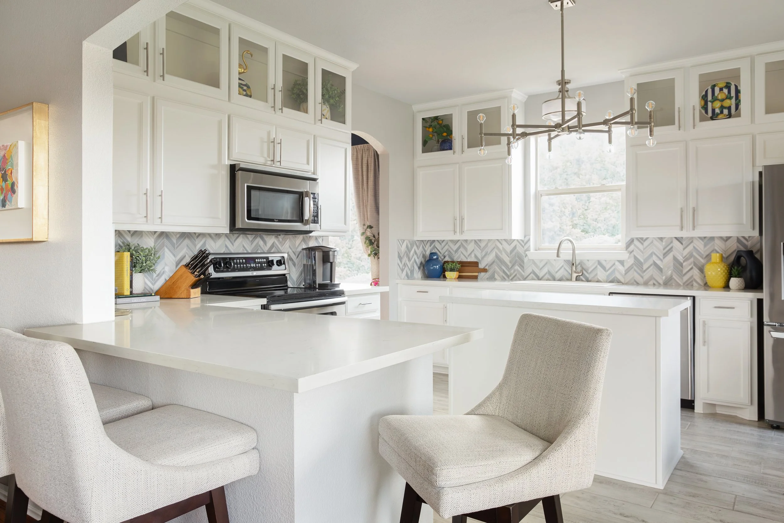 Modern kitchen with white cabinets, stainless steel appliances, and light gray chevron backsplash, featuring a white island with upholstered bar stools.
