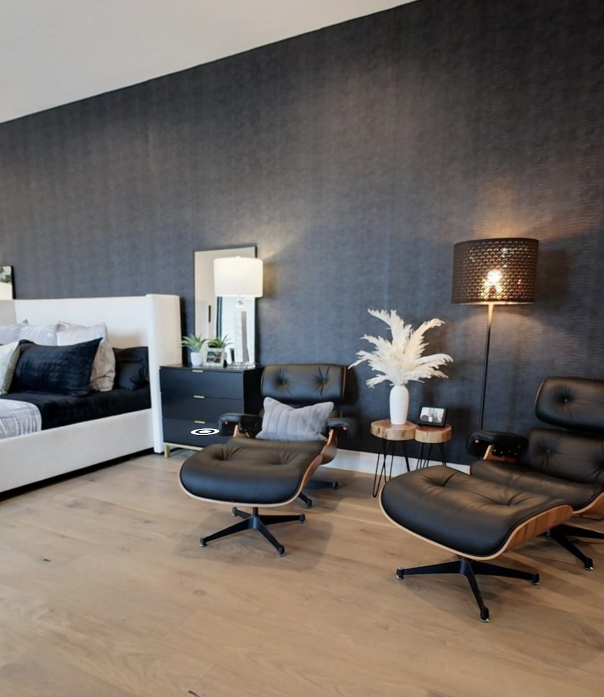 Modern bedroom with dark accent wall, featuring a white upholstered bed, black side table with lamp, two black leather lounge chairs, a small wooden table, and a floor lamp.