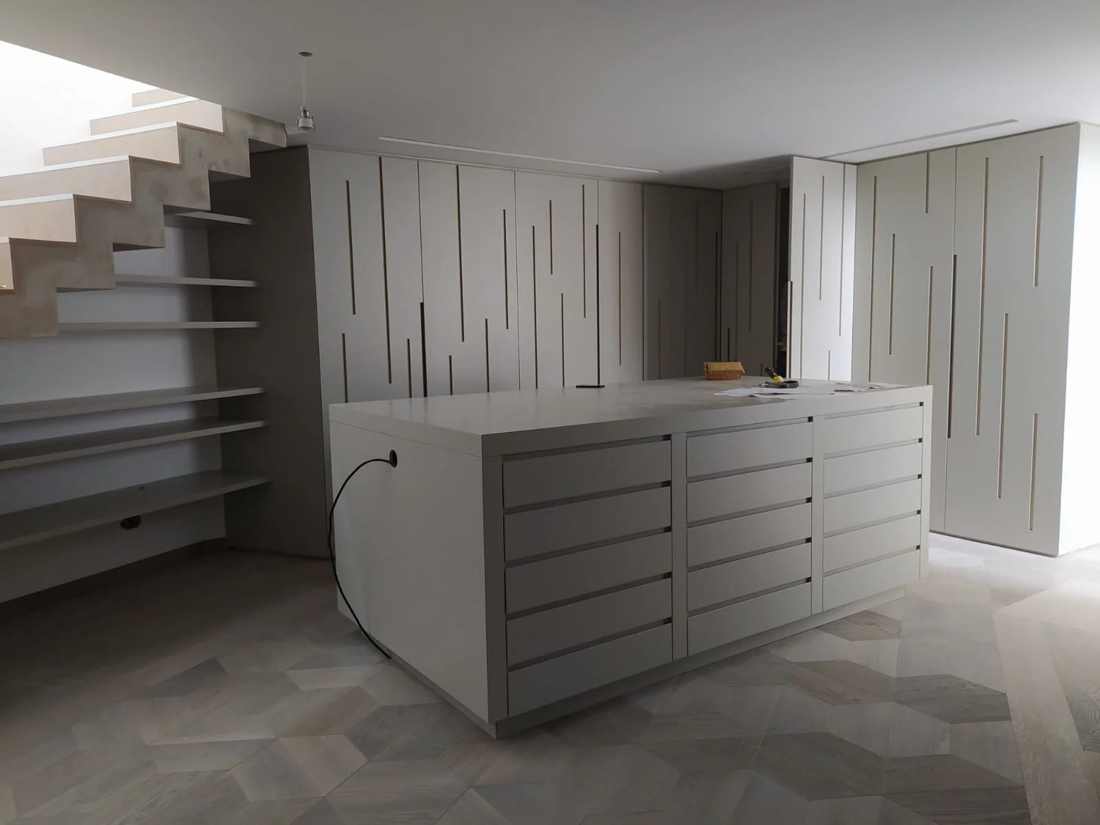 Cuisine moderne en cours de construction avec un îlot central blanc et un mur en panneaux ajustés. Étagères près de l'escalier et sol en carreaux de bois en chevrons.