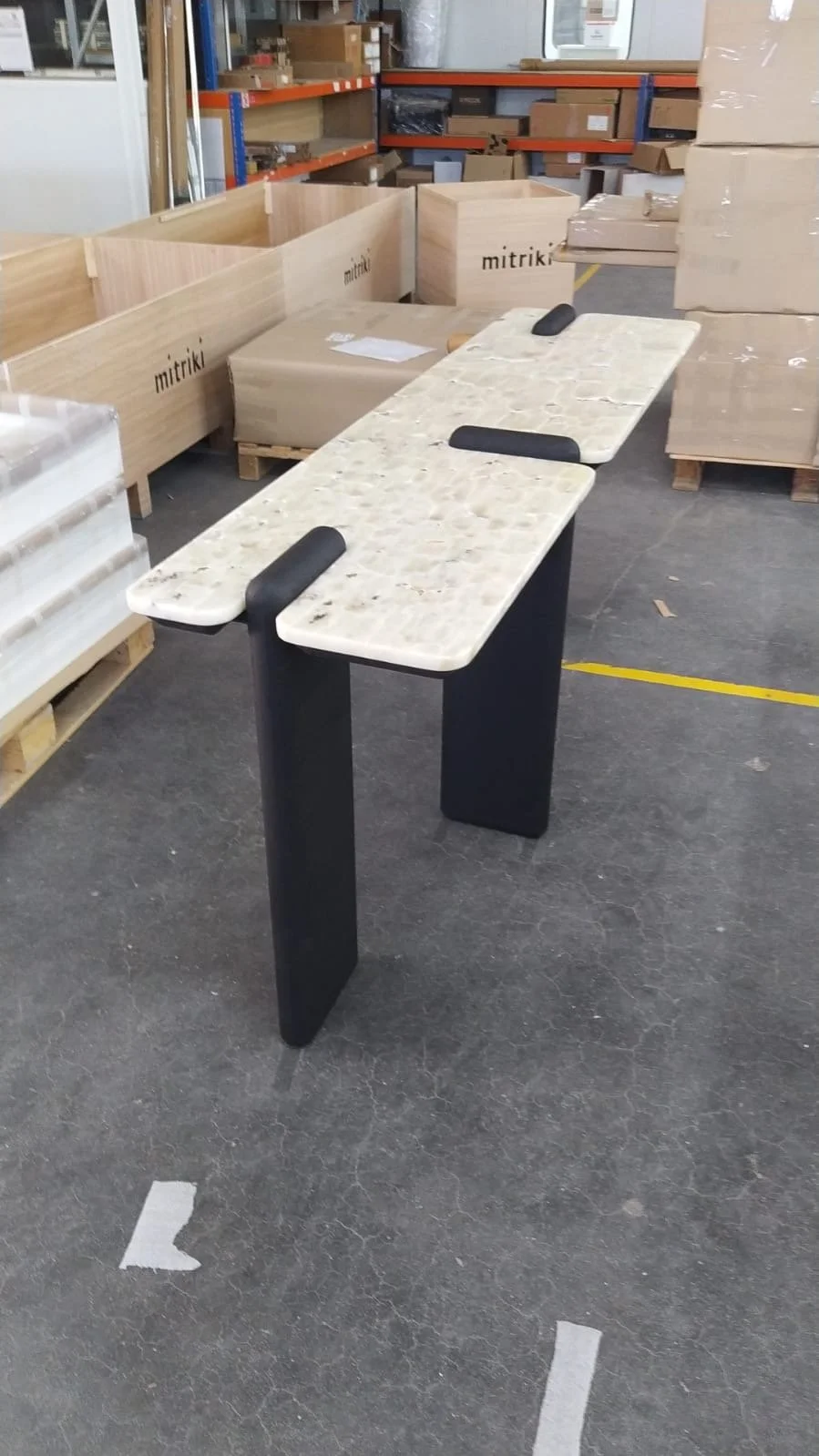 Une table avec un plateau en pierre ou marbre beige clair, pieds noirs, dans un entrepôt avec des cartons et des palettes autour.