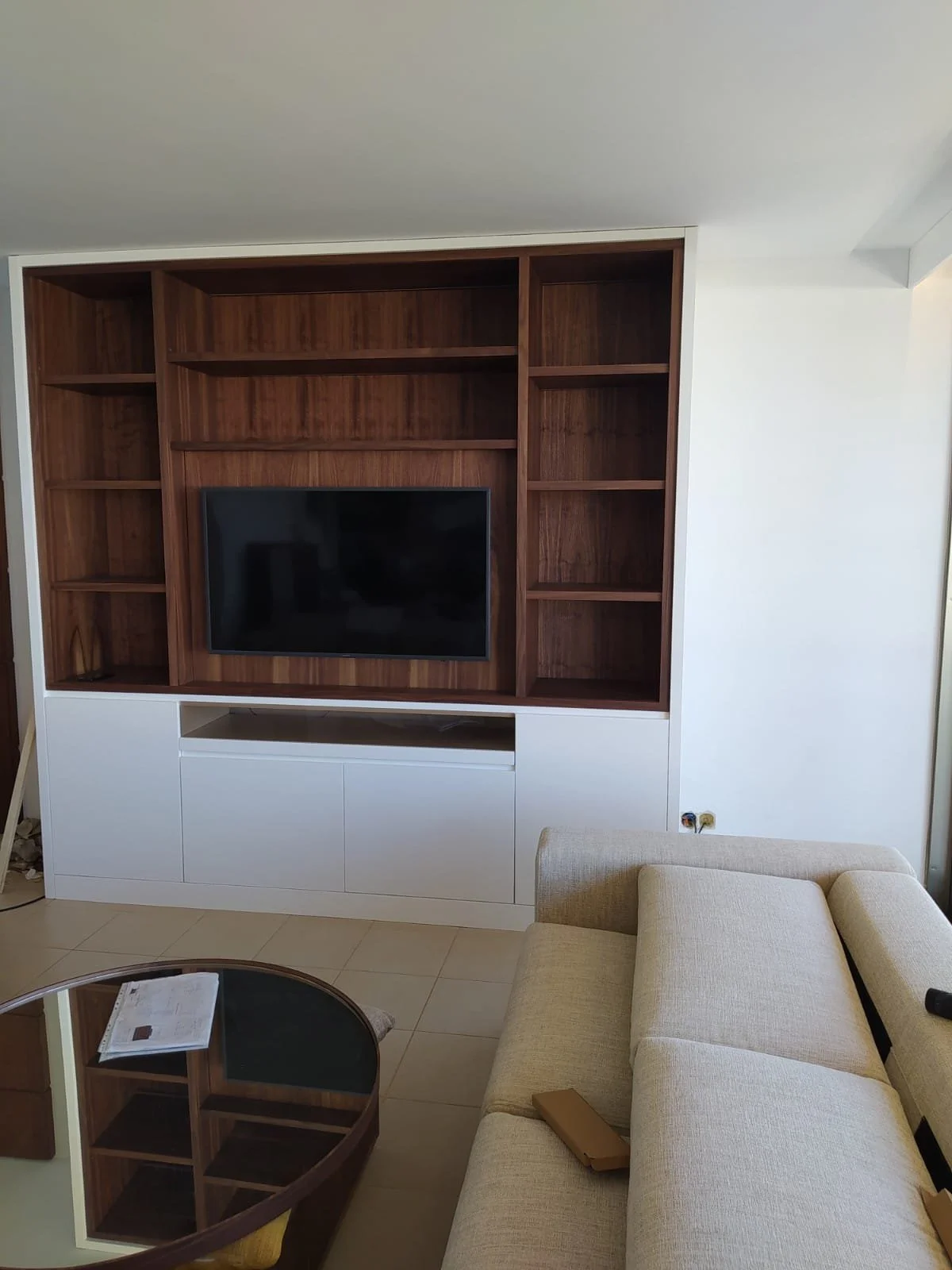 Salon avec un mur en bois et un téléviseur encastré, canapé beige, table en verre, murs blancs