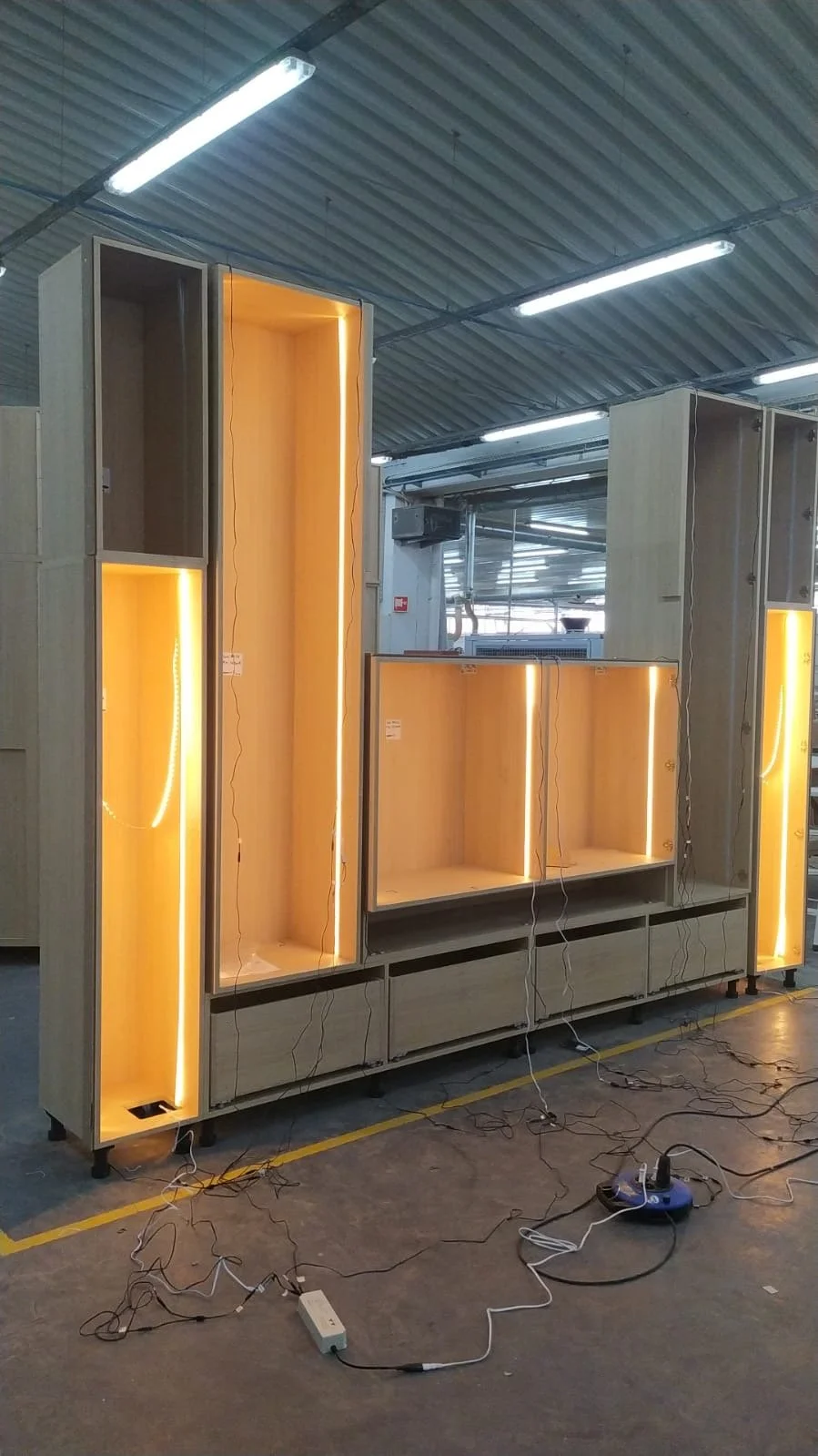 Étagères en bois avec éclairage led chaud dans un environnement d'usine ou atelier, certaines parties en câbles électriques suspendus ou connectés à un dispositif électrique sur le sol.