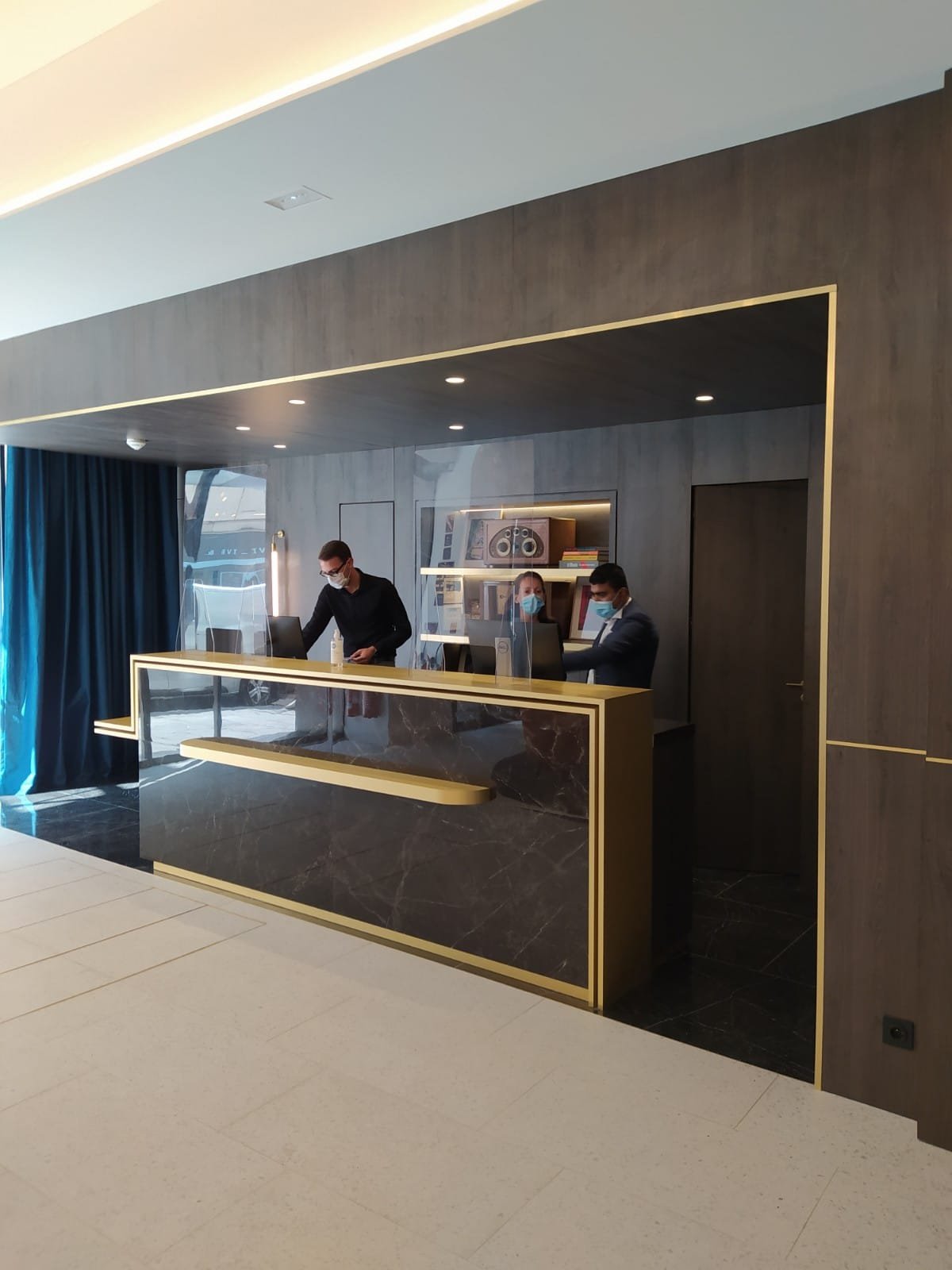 Une réception d'hôtel moderne avec trois employés portant des masques, travaillant derrière un comptoir en marbre noir avec des détails dorés.