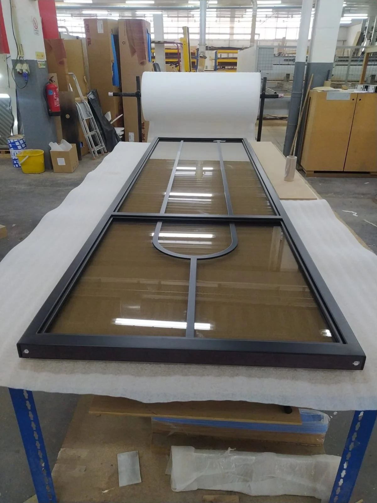 Une grande fenêtre en verre avec un cadre métallique posé sur une table dans un atelier. La fenêtre semble destinée à une installation extérieure ou un bâtiment.