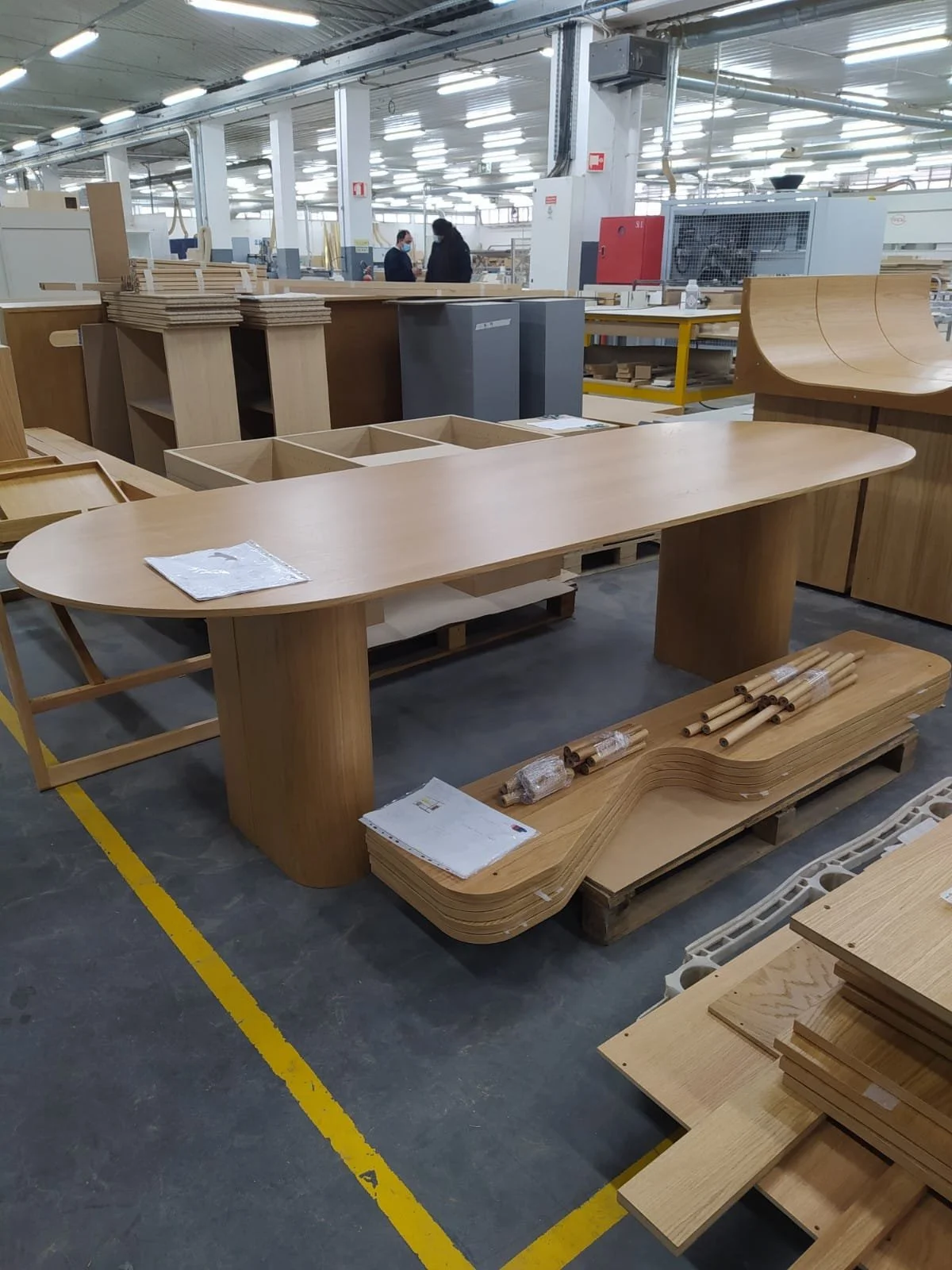 Table en bois en cours de fabrication dans une usine, avec d'autres meubles en bois et des employés en arrière-plan.