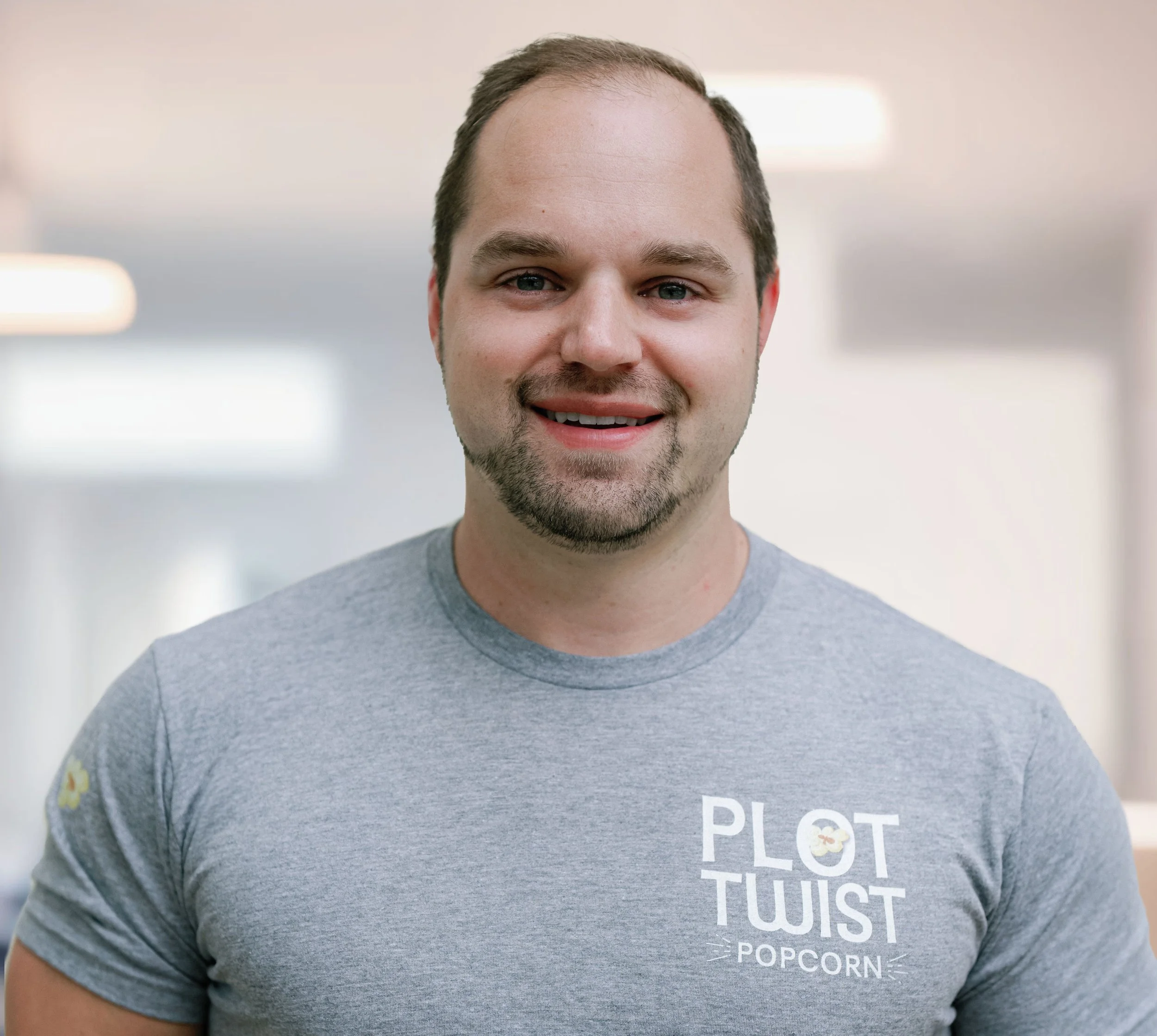 Smiling man wearing a gray T-shirt with 'Plot Twist Popcorn' logo, standing in an indoor setting.