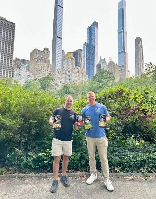 Two men standing outdoors in Central Park, New York City, holding books titled 'Plot Twist', with skyscrapers in the background.
