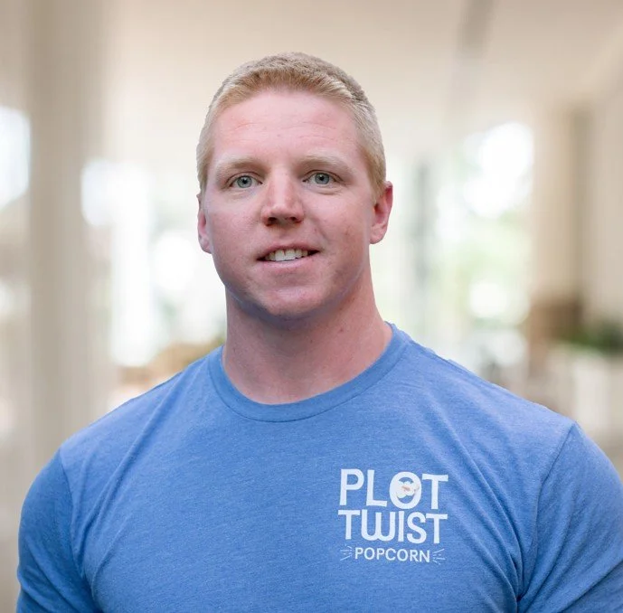 A young man with short blond hair wearing a blue t-shirt that says 'Plot Twist Popcorn,' standing indoors with a blurred background.