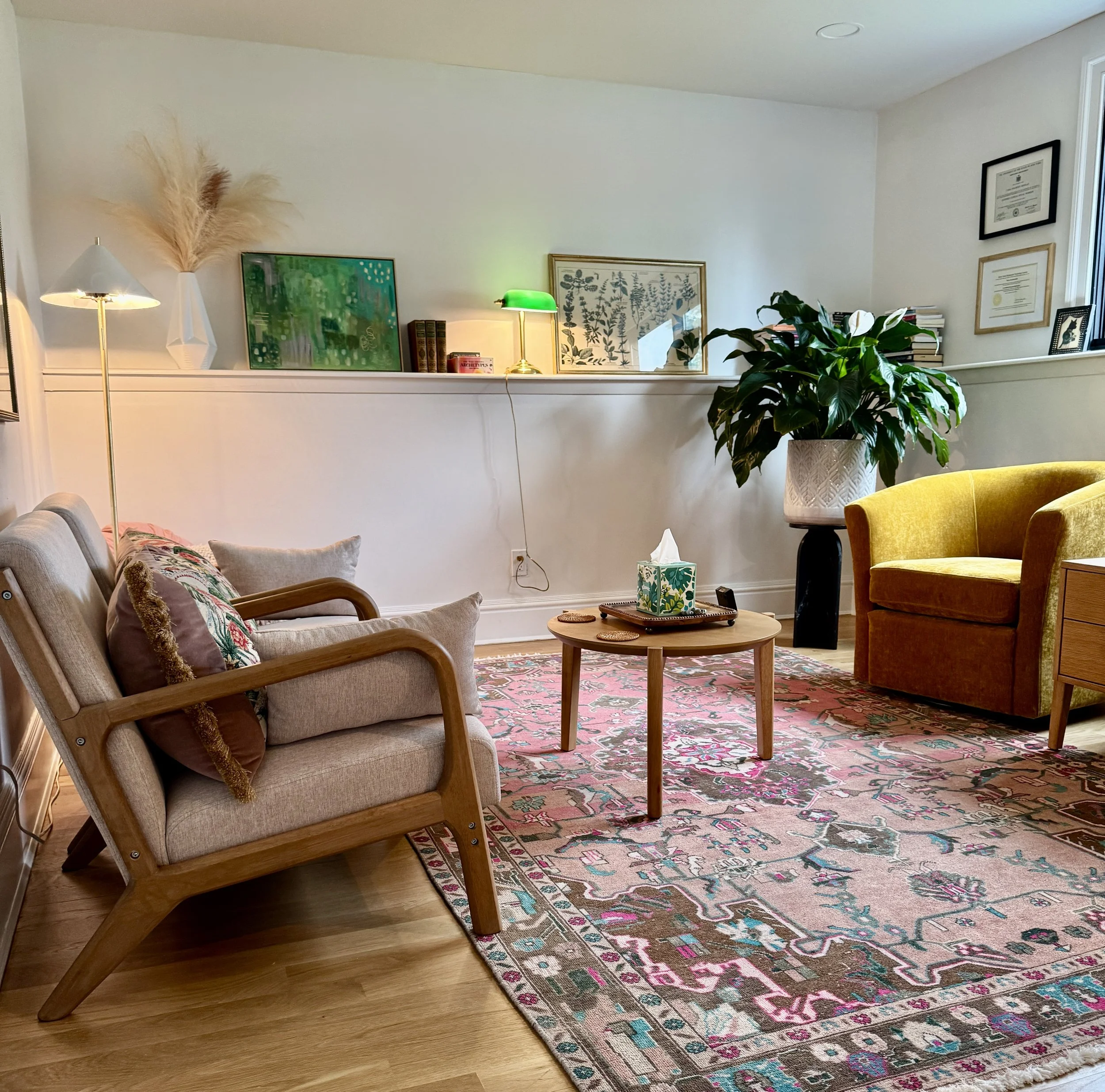 Living room with a patterned pink rug, beige armchair, green armchair, large plant, books, and framed pictures on the wall.