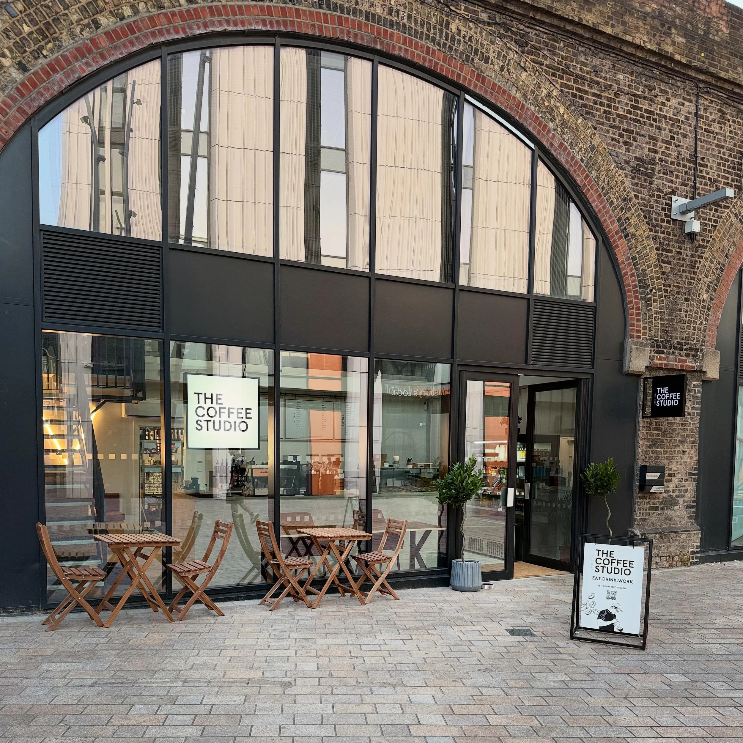 Front view of a coffee shop named 'The Coffee Studio' with outdoor seating, including three wooden tables and six chairs, and a black signboard with the shop's name and slogan, set on a brick sidewalk outside a building with large glass windows and brick archways.