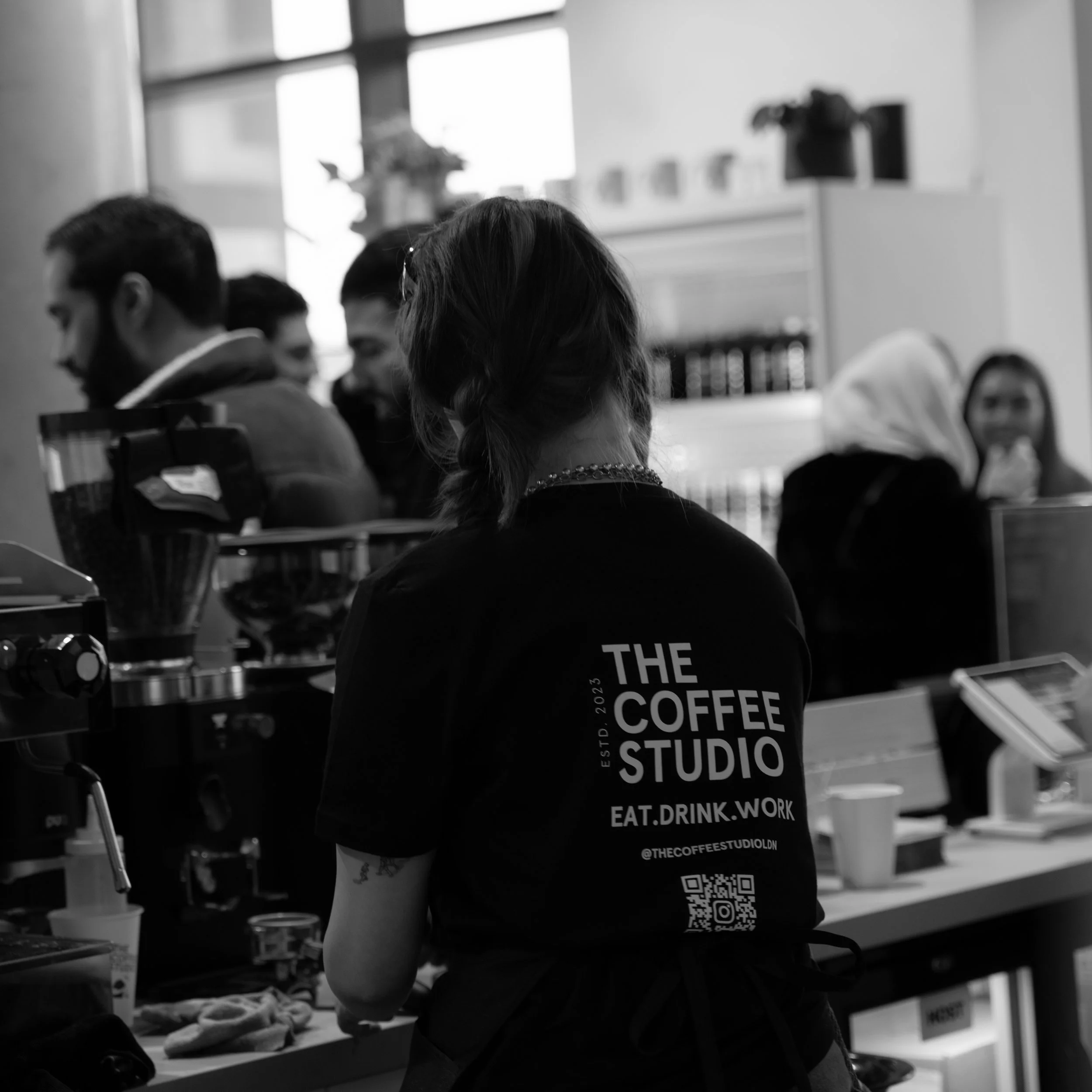 A woman with a braid and a tattoo on her arm stands behind a coffee counter at The Coffee Studio, surrounded by baristas and customers in a busy cafe. She is wearing a black apron with the cafe's logo and text.