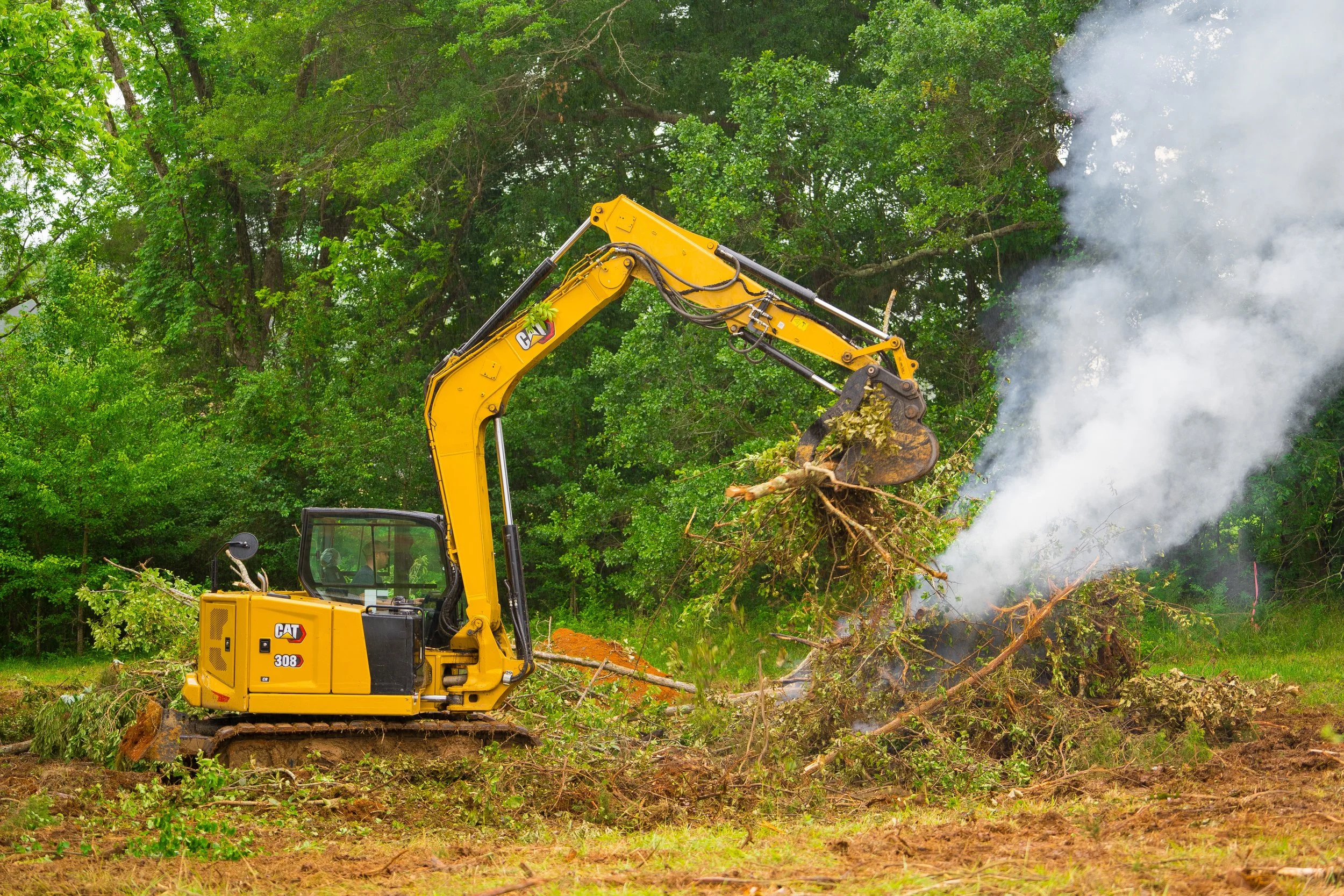 A yellow Caterpillar excavator is uprooting trees in a forested area; steam or smoke is rising from the scene.